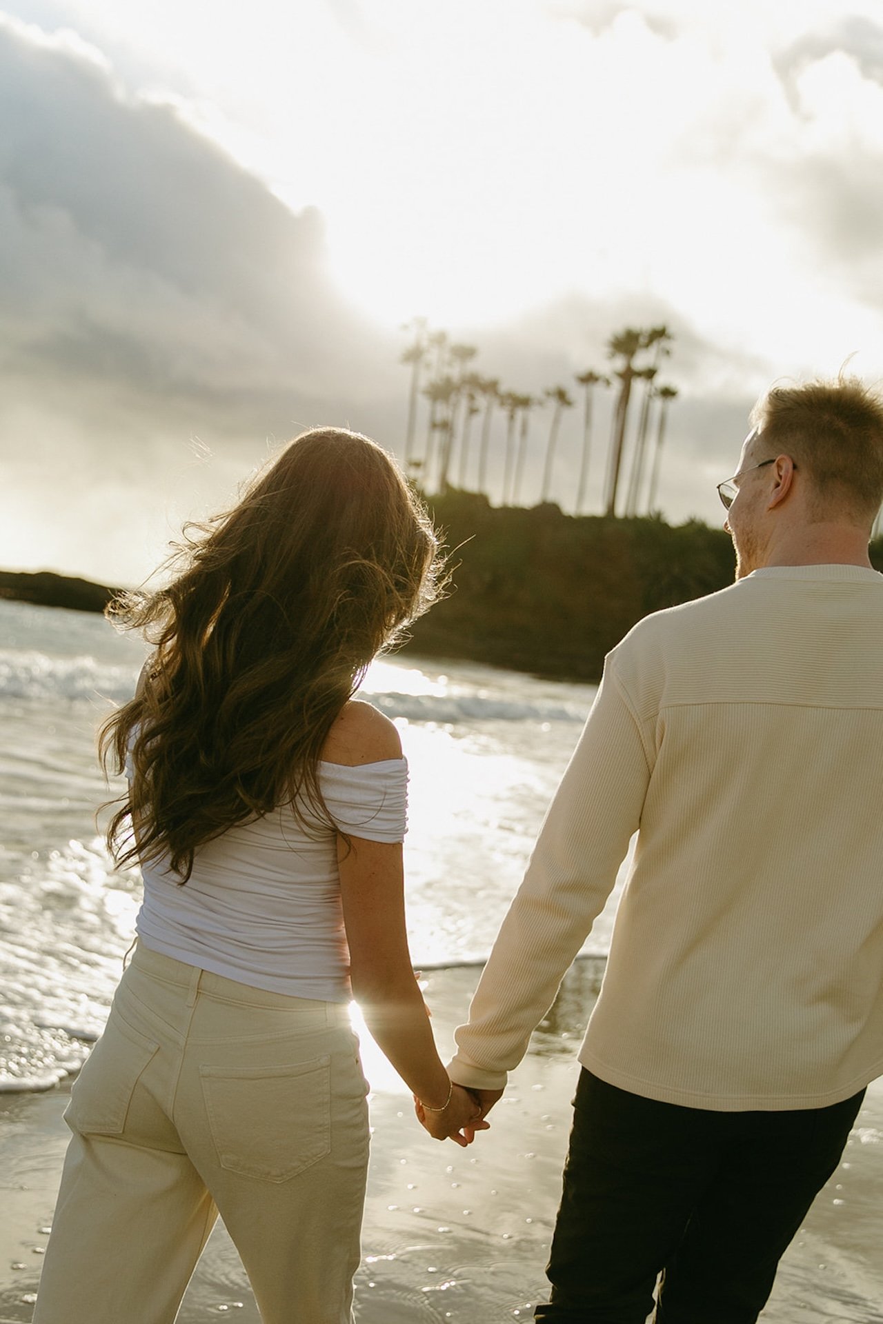 A couple holding hands while walking along the shoreline, backlit by golden sunset light with palm trees silhouetted against the sky.
