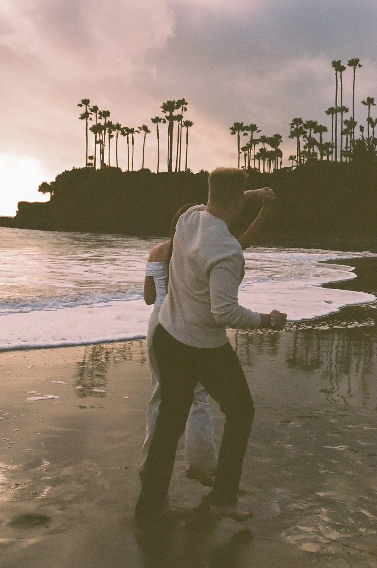 A couple playfully moving through shallow water at sunset, creating soft motion blur with reflections in the wet sand and pastel sky overhead.