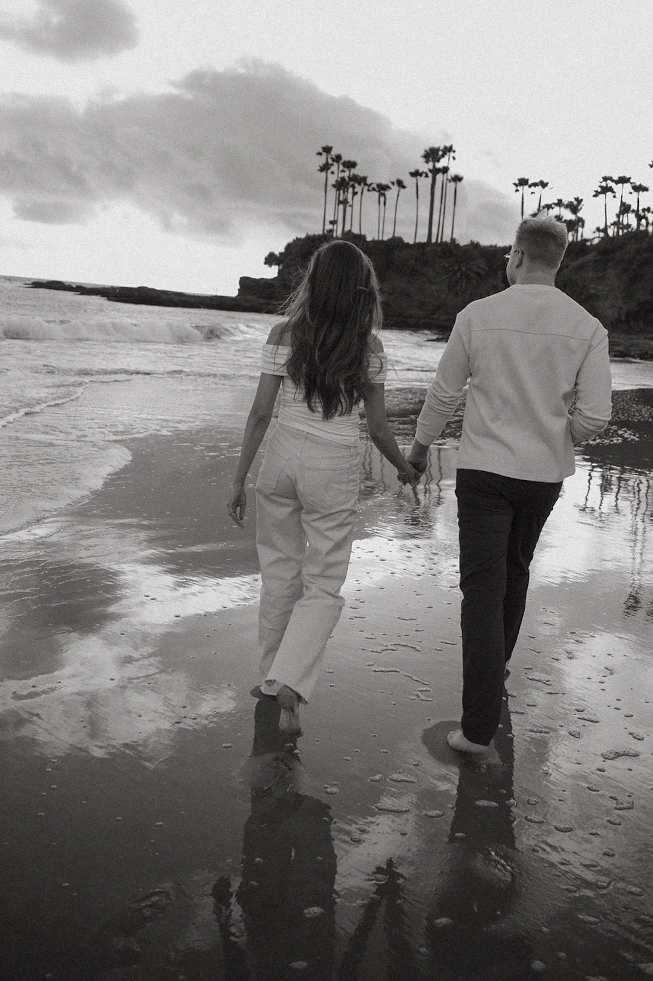 Black and white image of a couple walking hand in hand along the beach, ocean waves and palm trees creating a moody coastal backdrop.