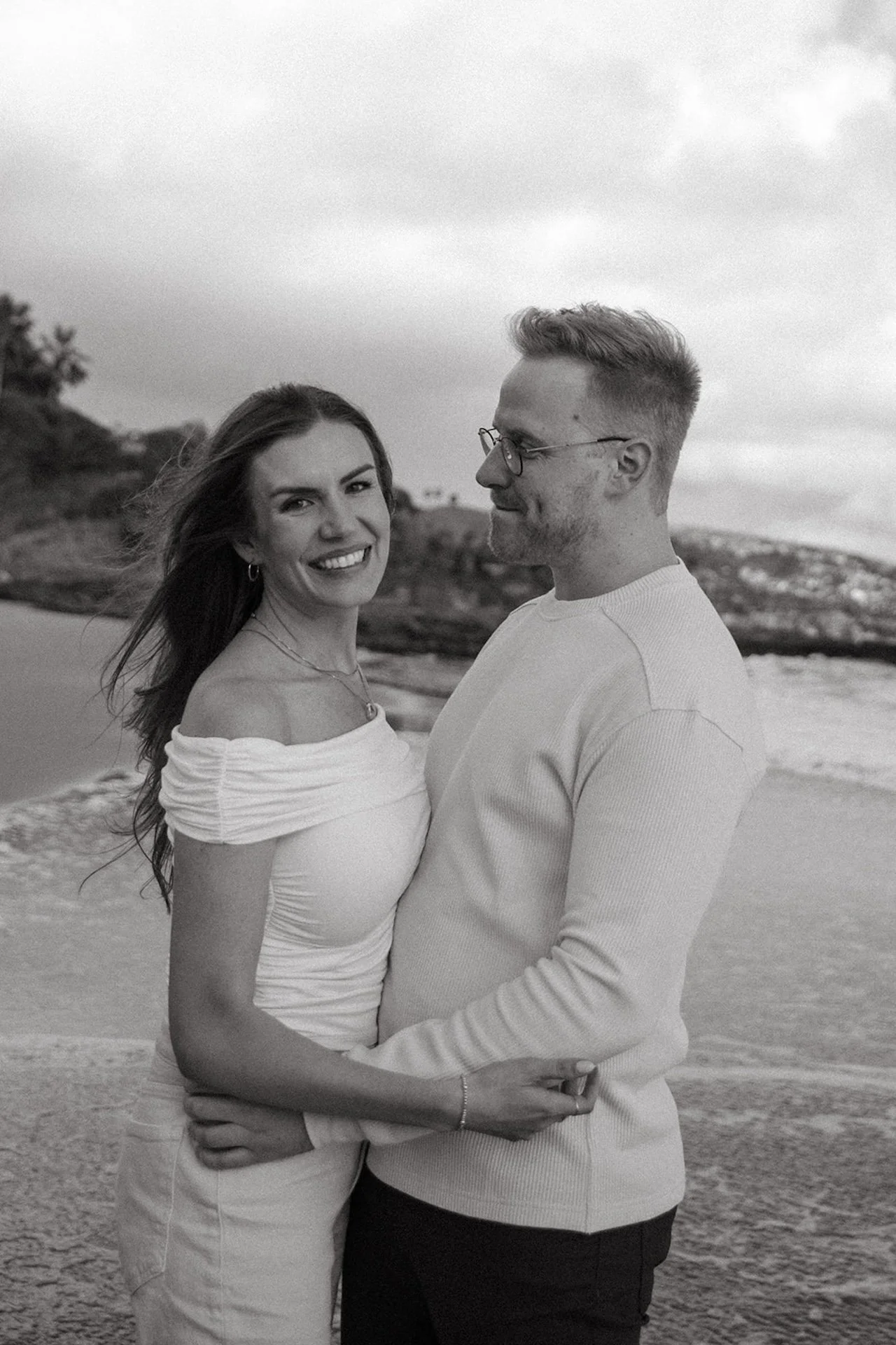 Black and white portrait of a couple standing close together on the beach, smiling at each other with the ocean and shoreline behind them.
