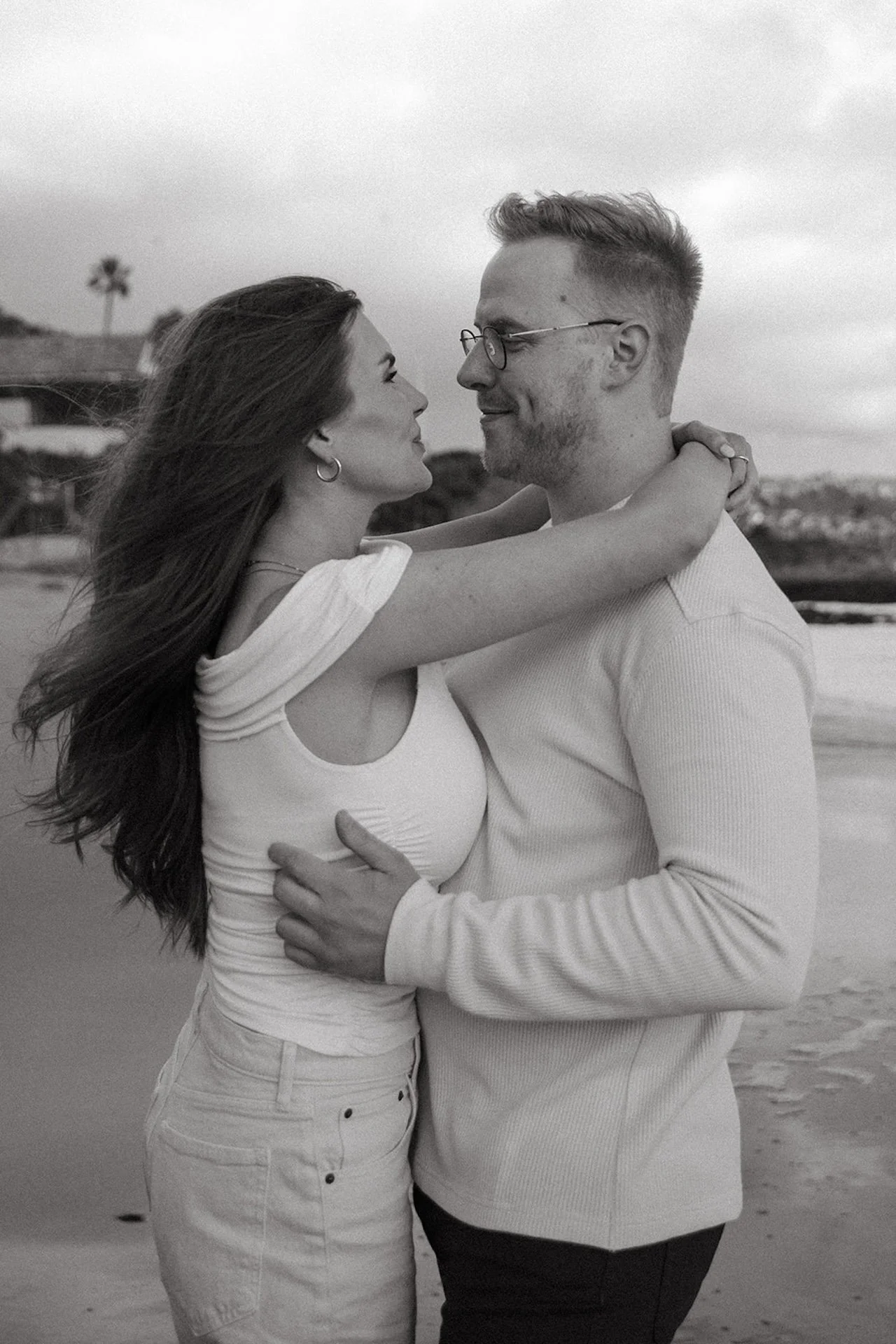 Black and white portrait of a couple standing close together on the sand, arms wrapped around each other and smiling softly during laguna beach engagement photos at shaws cove laguna beach.