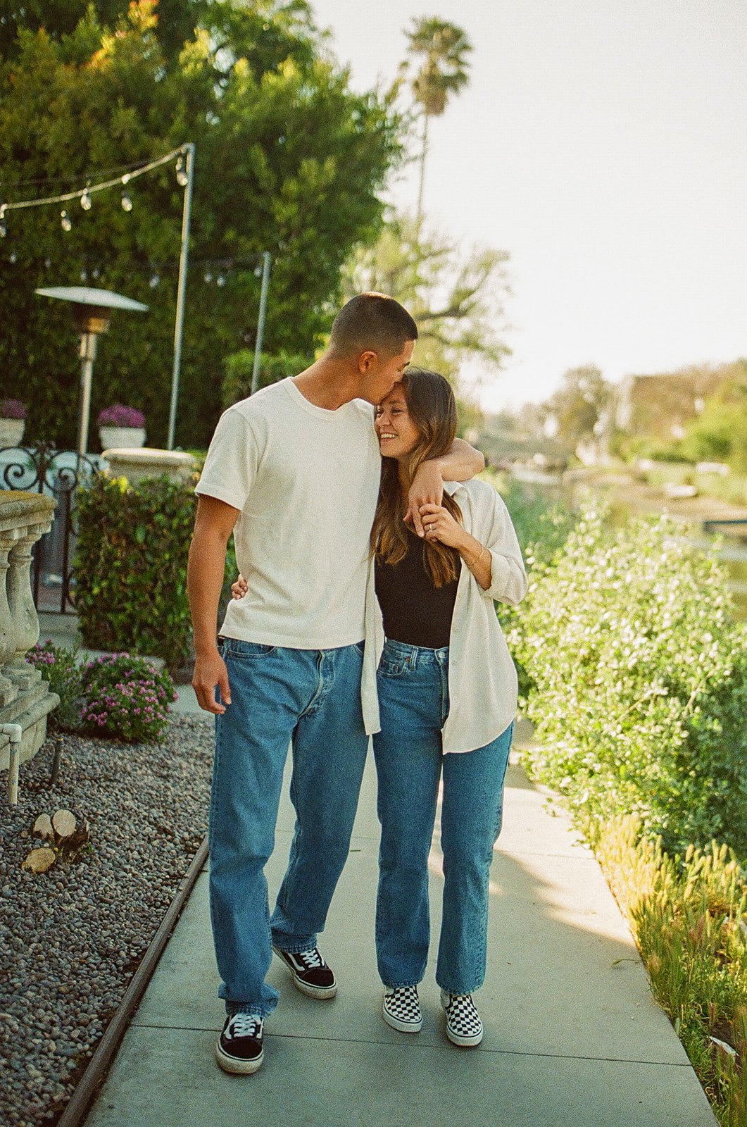 A couple walking arm in arm down a quiet Venice neighborhood sidewalk surrounded by greenery during golden hour.