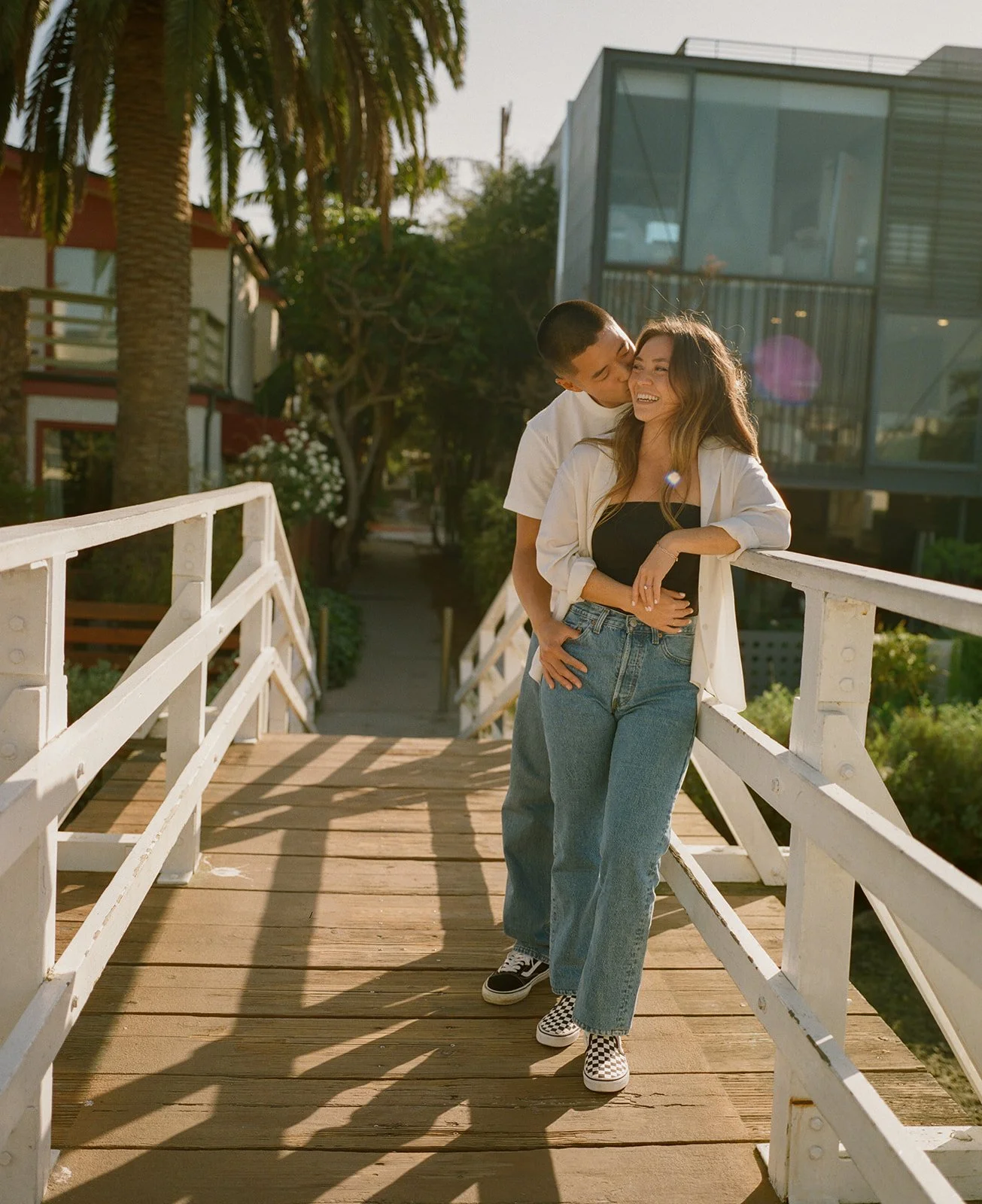 A couple standing on a bridge as the man wraps his arms around his fiance with palm trees behind them