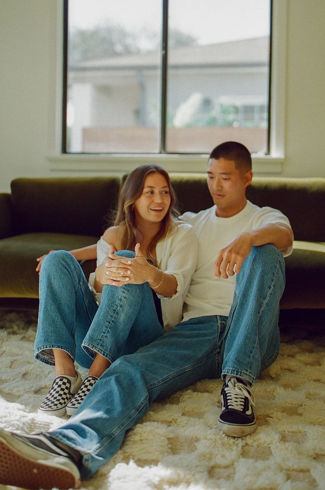 A couple sitting on the floor leaning into each other in front of a window during a casual at-home session.