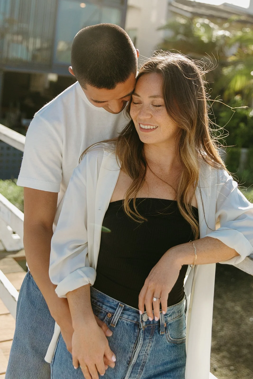 A close-up portrait of a couple standing together on a white bridge with greenery and water behind them.
