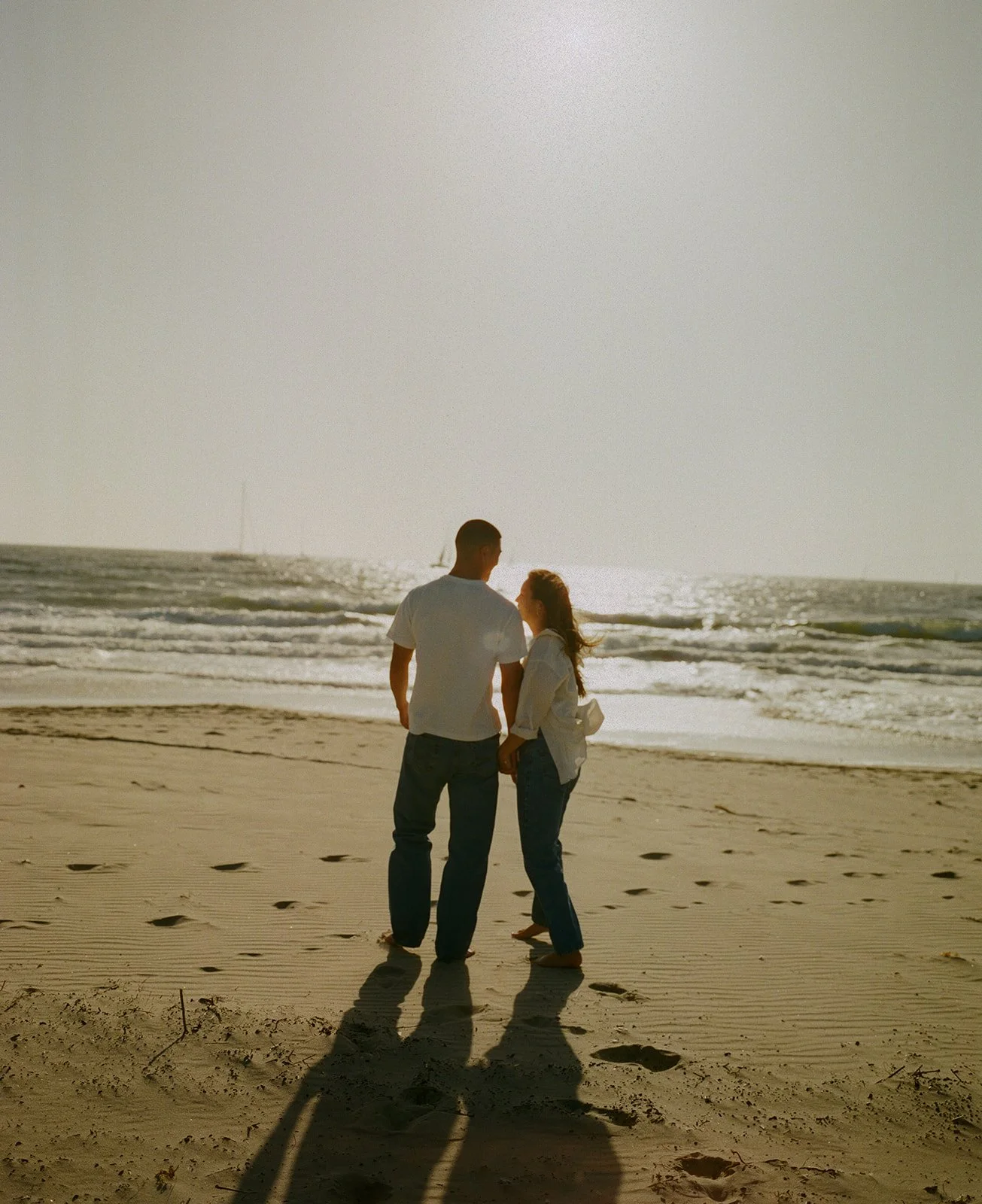 A couple standing barefoot on the sand facing each other as the ocean glows behind them.