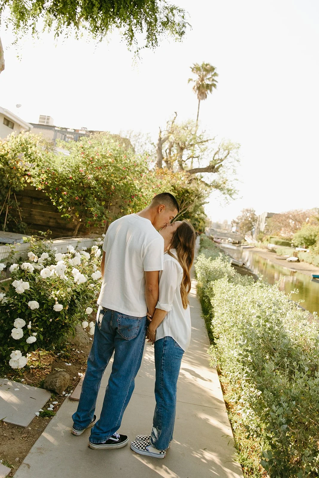 A quiet neighborhood moment of a couple kissing on a pathway, captured at Venice Beach Photoshoot Locations with palm trees, coastal homes, and a relaxed street scene.