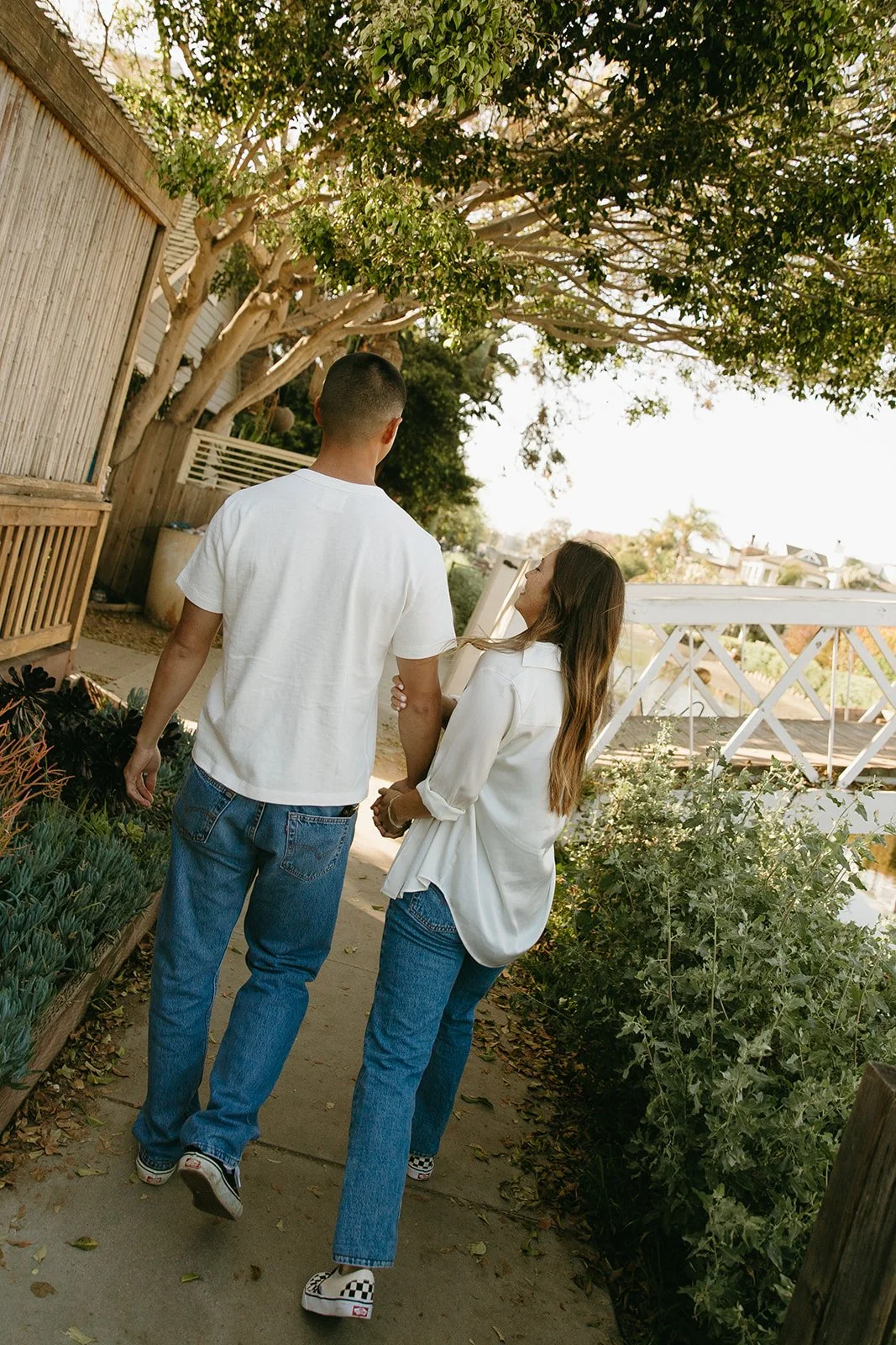 A couple walking hand in hand down a leafy path surrounded by greenery and warm afternoon light.