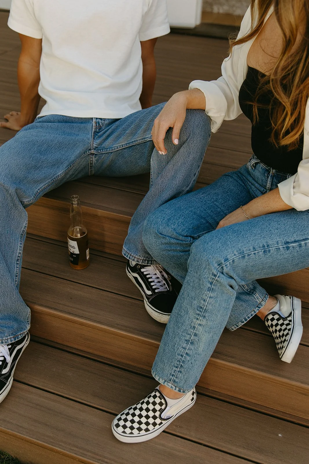 A close-up detail of a couple sitting on wooden steps in casual denim with a beer bottle resting between them.