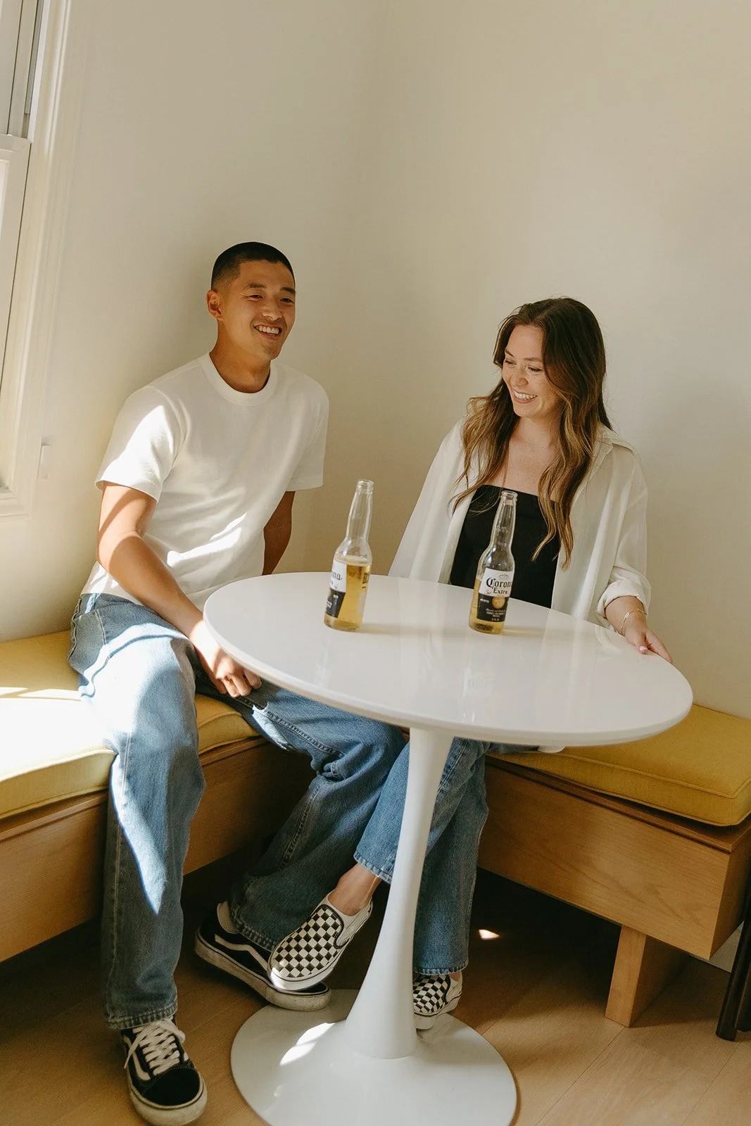 A couple laughing at their kitchen table, enjoying a beer, during their at home engagement session