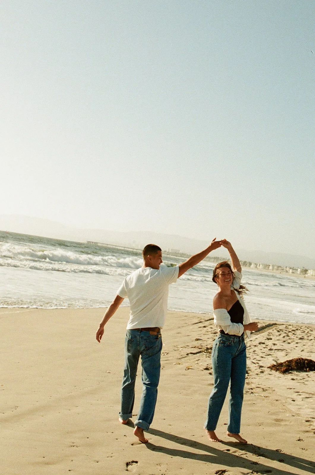 A playful moment of a couple spinning and laughing barefoot on the sand near the shoreline.