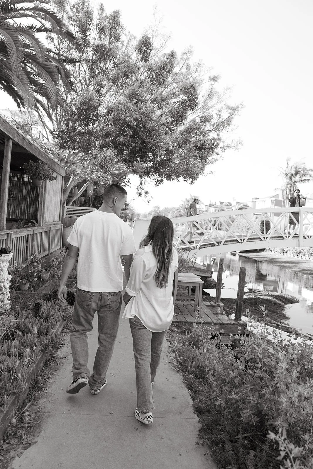 A black and white image of a couple walking side by side along a canal path with greenery and a small bridge ahead.