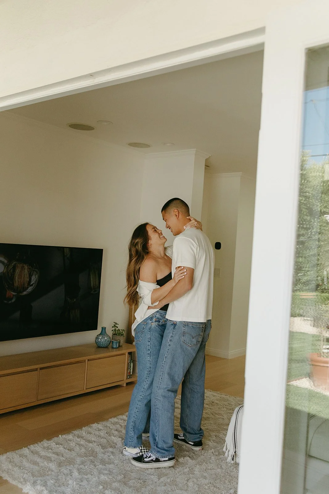 A couple standing close together in their living room, smiling and holding each other in a relaxed at-home moment.