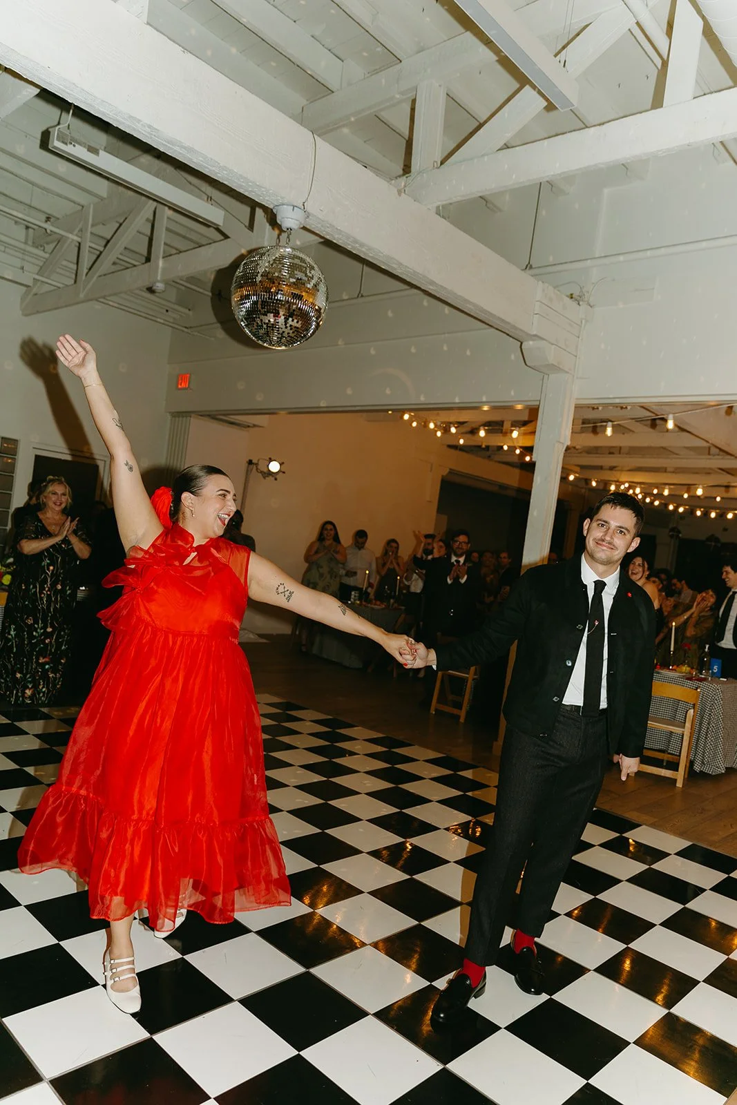 The couple making a joyful entrance onto a black and white checkered dance floor at The Revery LA as guests cheer in the background.