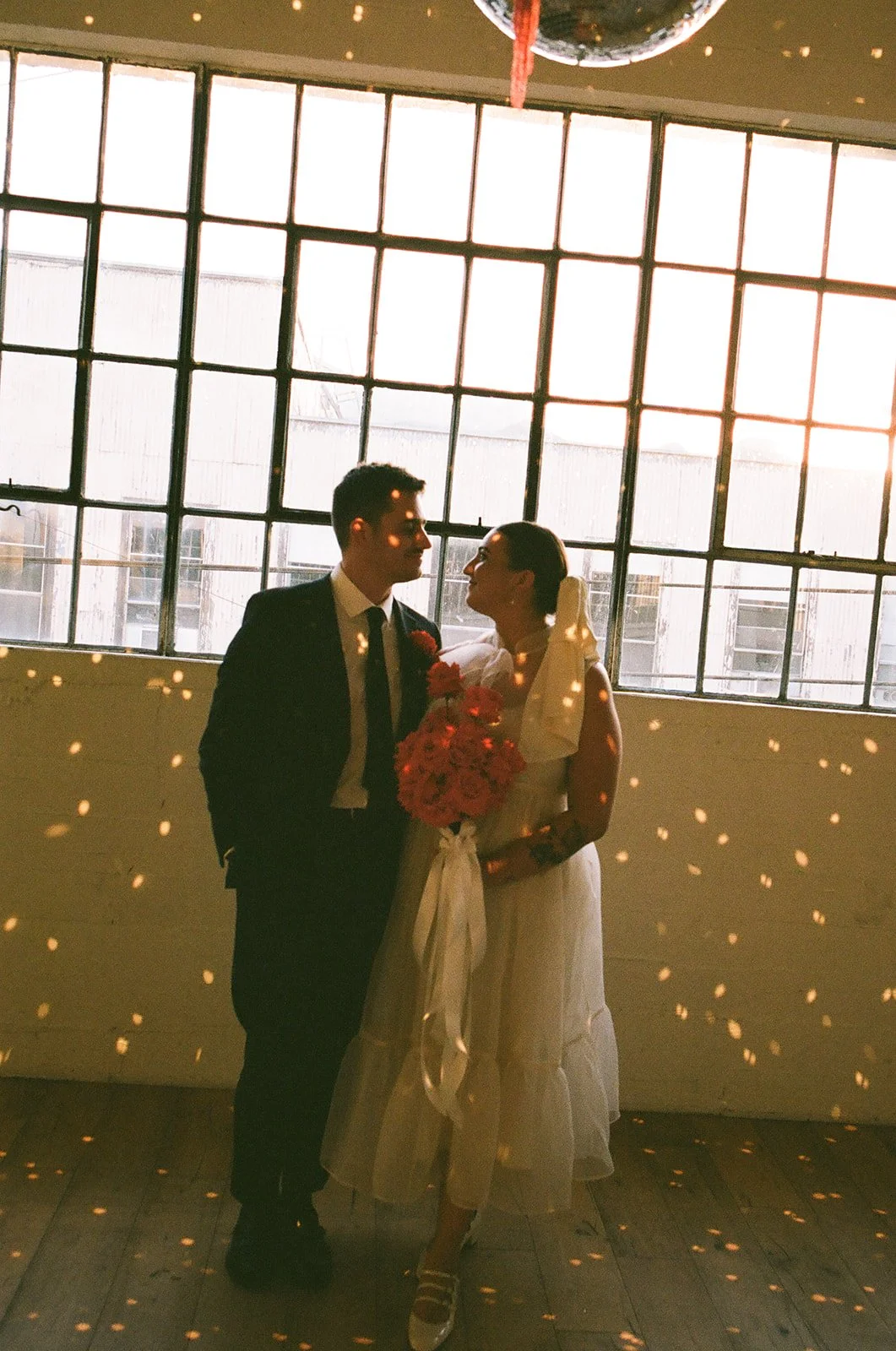 The couple standing together in front of large windows at The Revery LA, surrounded by disco ball reflections and soft evening light.