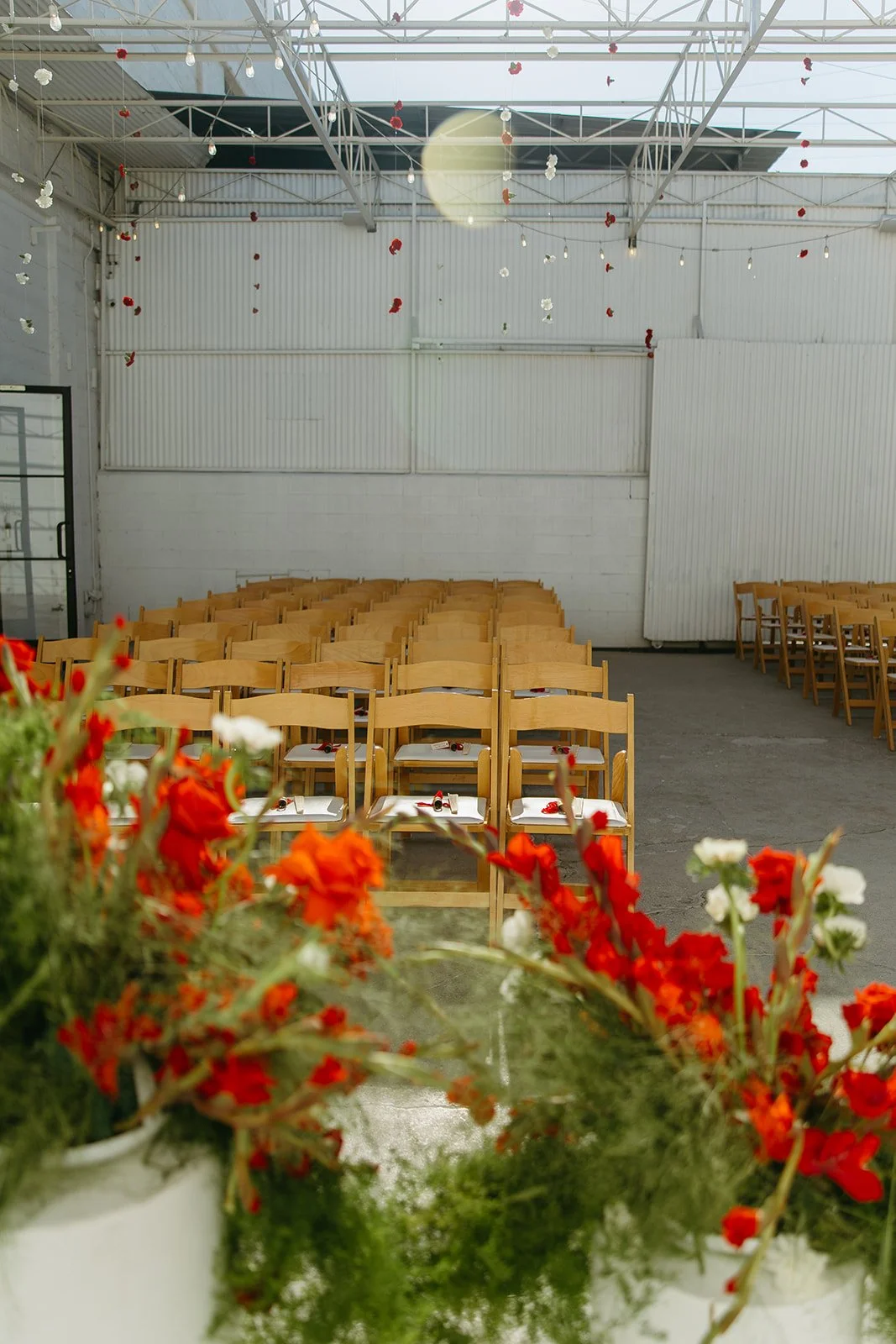 Rows of wooden ceremony chairs set up inside The Revery LA with red florals lining the aisle and soft light coming through the ceiling.