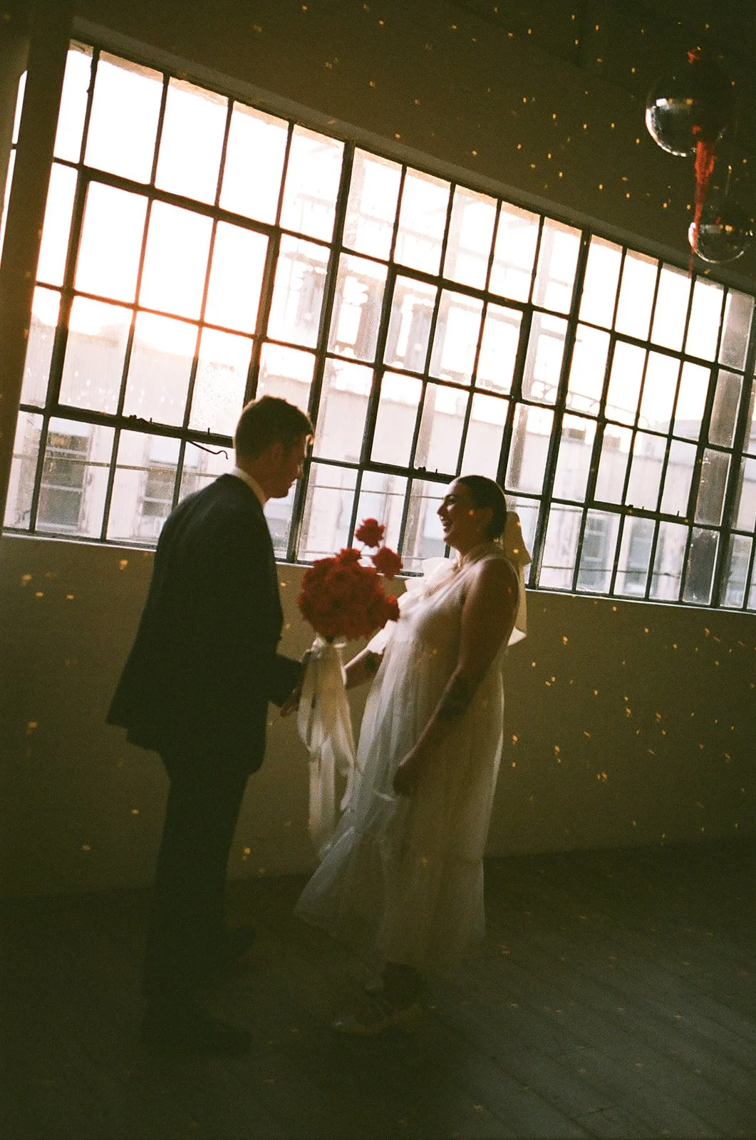 A soft candid film moment of the bride and groom standing in front of a huge window pane with the sunsetting behind them