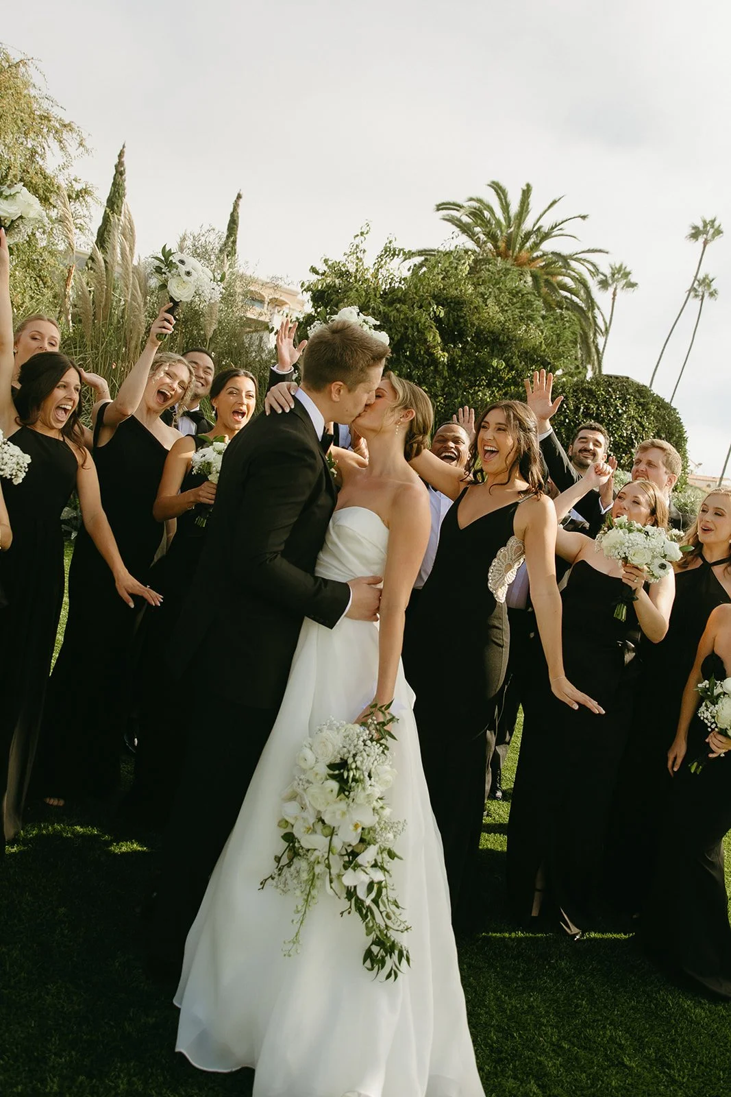 Bride and groom kissing in front of their wedding party during an outdoor celebration at one of the most iconic San Clemente Wedding Venues, surrounded by palm trees and coastal light.