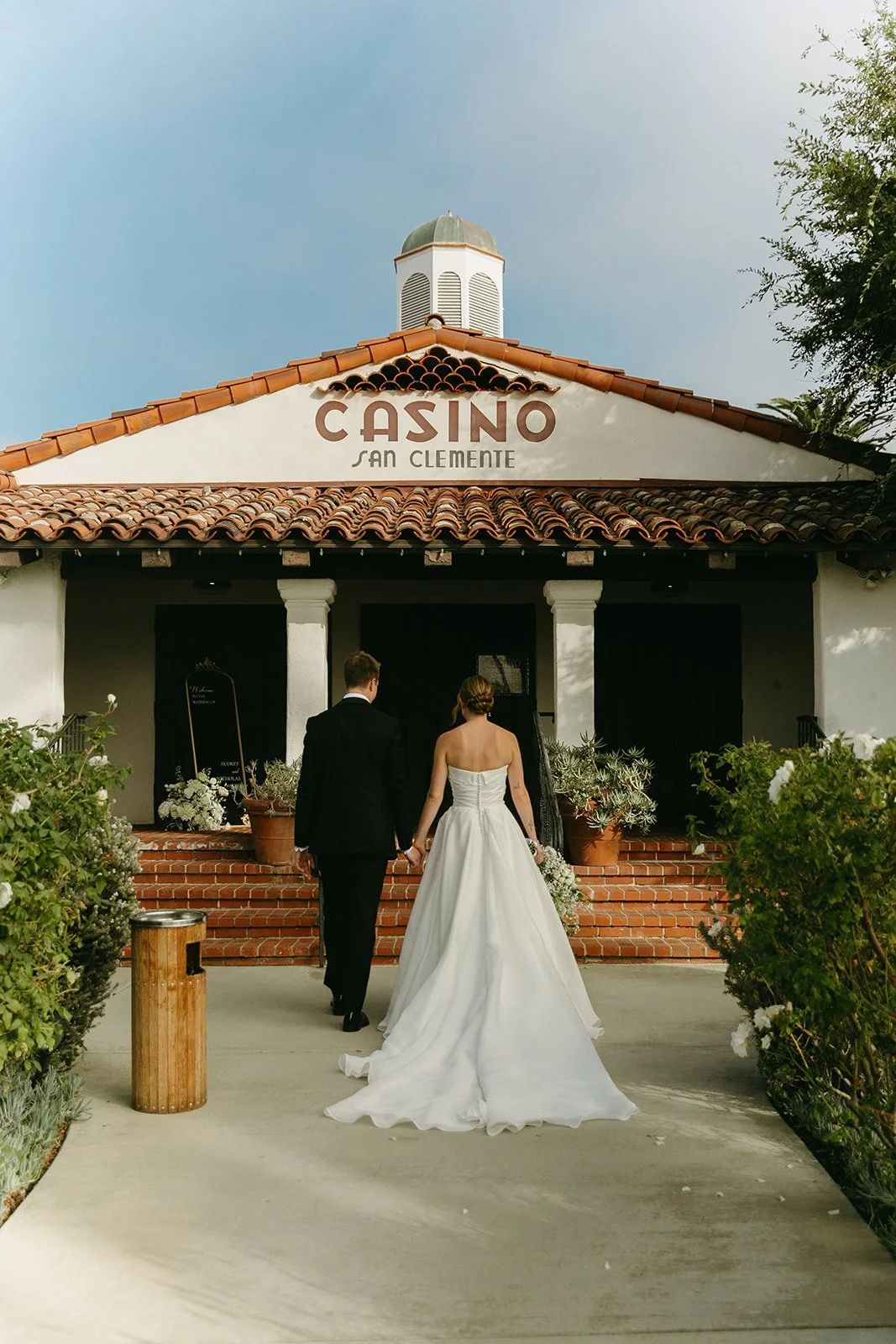 Bride and groom walking hand in hand toward the Casino San Clemente building, showcasing Spanish-style architecture and terracotta roof details.