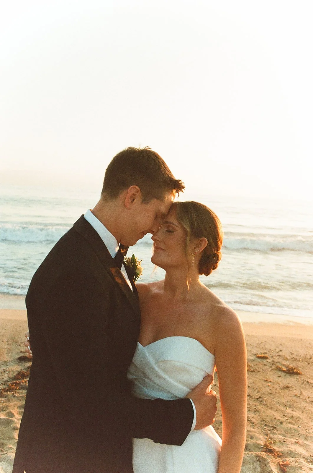 Intimate coastal portrait of the newlyweds standing close together on the shoreline at sunset.