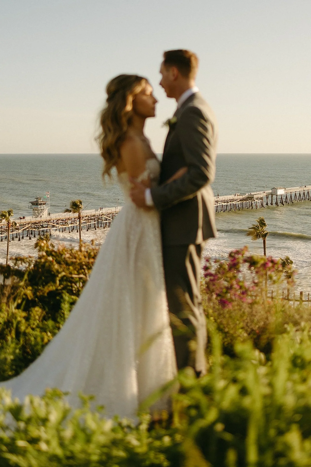 Soft-focus portrait of the bride and groom embracing with the San Clemente Pier in the background at one of the most romantic San Clemente Wedding Venues.