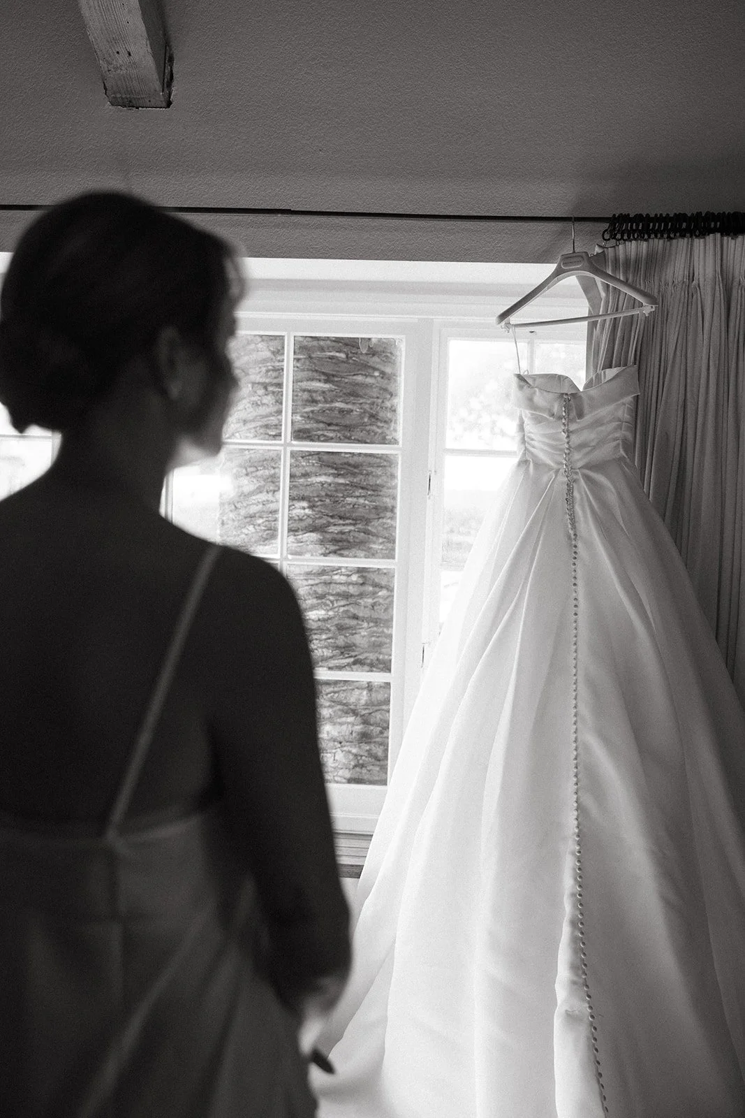 Bride standing in silhouette while looking at her wedding dress hanging in front of a window at one of the most timeless San Clemente Wedding Venues.