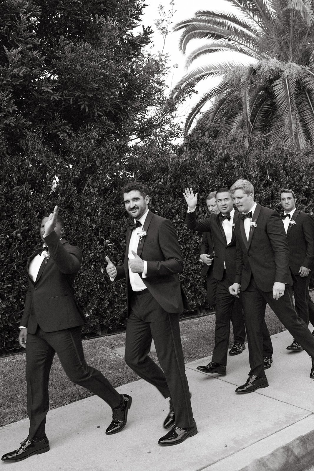 Black and white photo of groomsmen walking together in tuxedos, waving and smiling as they head toward the ceremony.