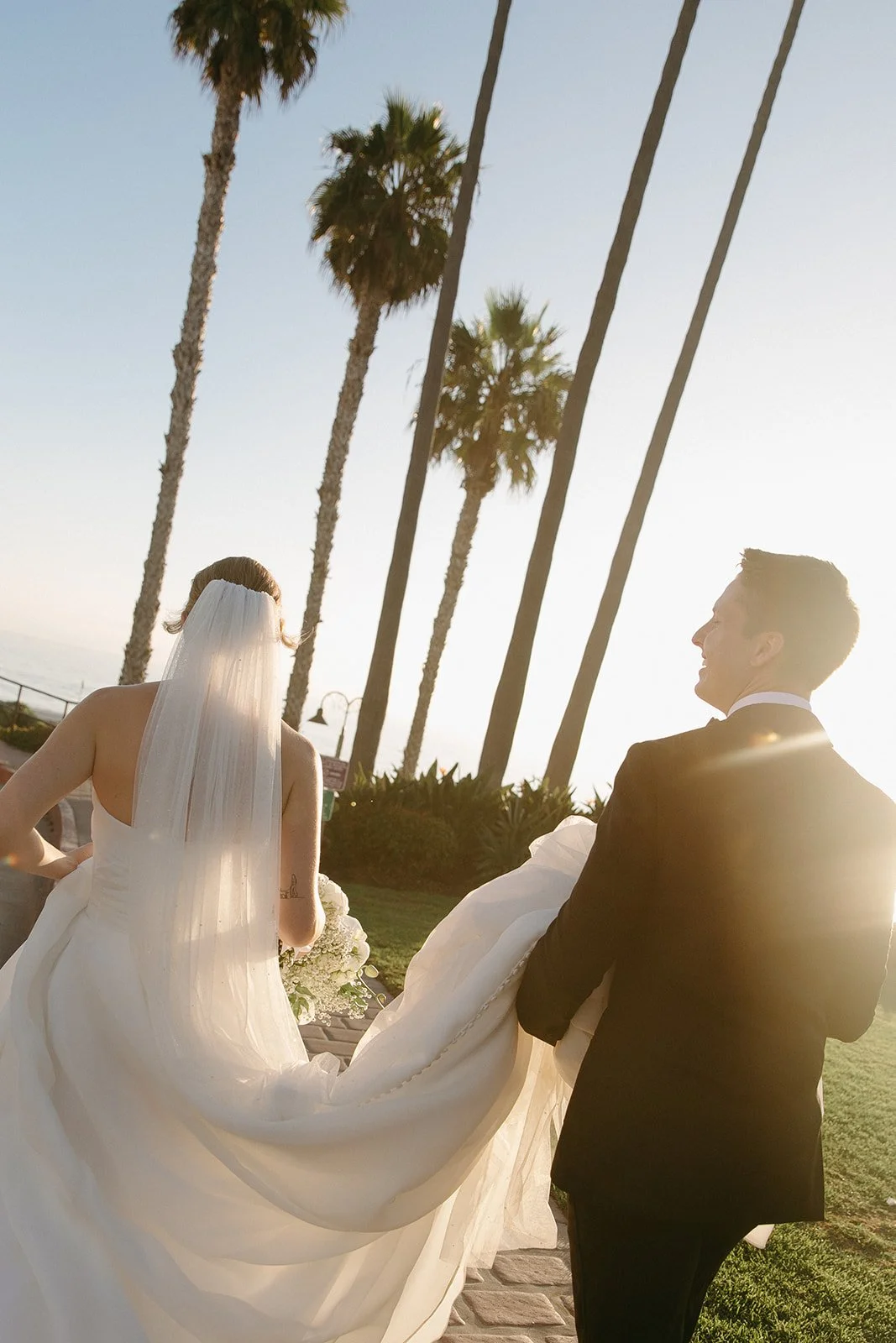 Candid photo of the couple laughing together as they walk toward the reception space, with the groom carrying the train of the wedding dress.