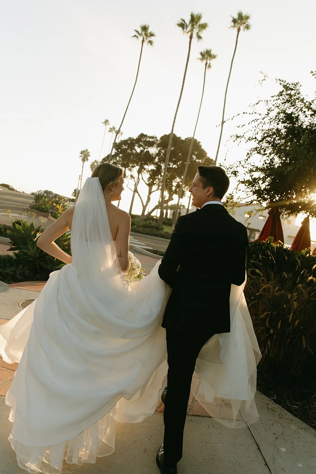 Bride and groom walking together through a palm-lined path at one of the most recognizable San Clemente Wedding Venues, with warm sunlight filtering through the trees.