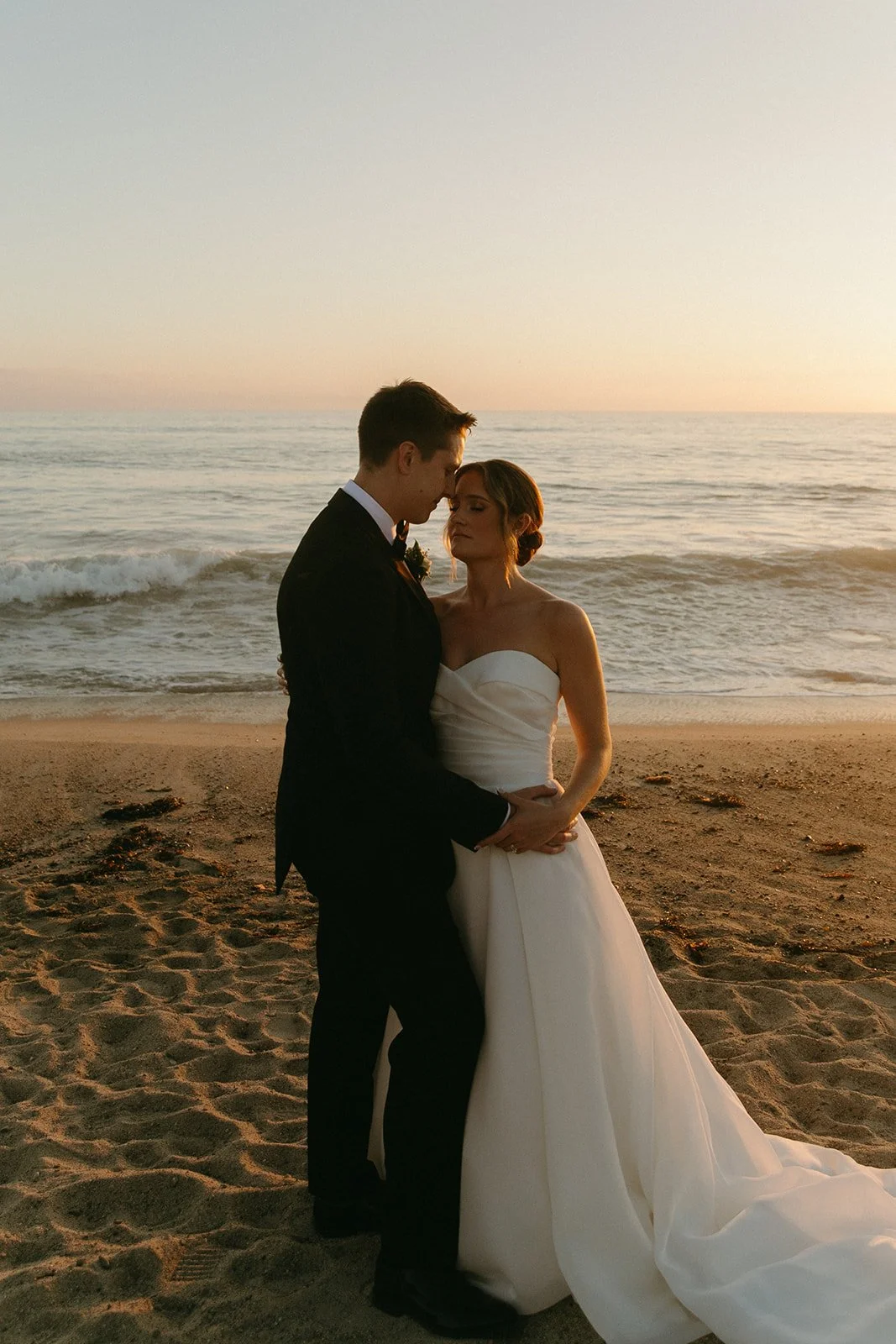Romantic sunset portrait of the bride and groom embracing on the beach near iconic San Clemente Wedding Venues, framed by soft waves and pastel sky tones.