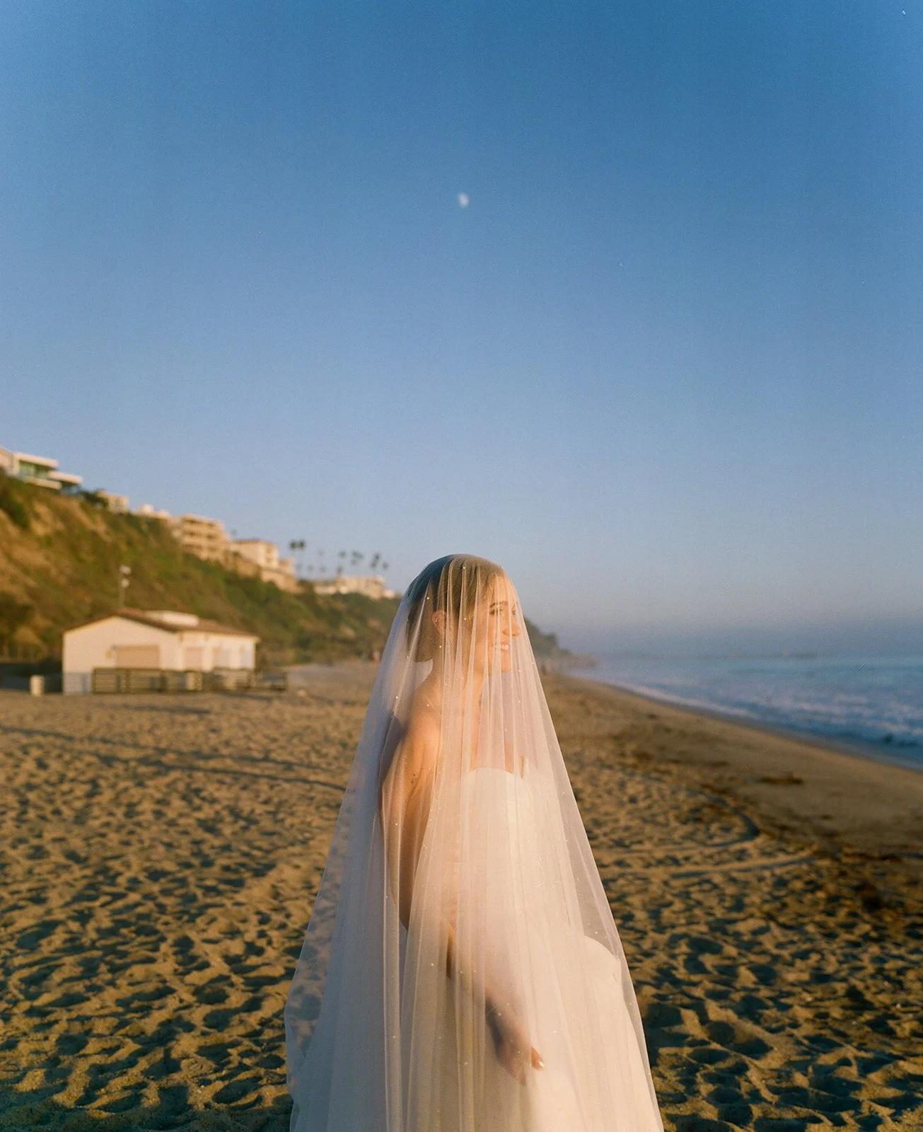 Bride standing alone on the beach at golden hour with her veil blowing in the ocean breeze near one of the most scenic San Clemente Wedding Venues.