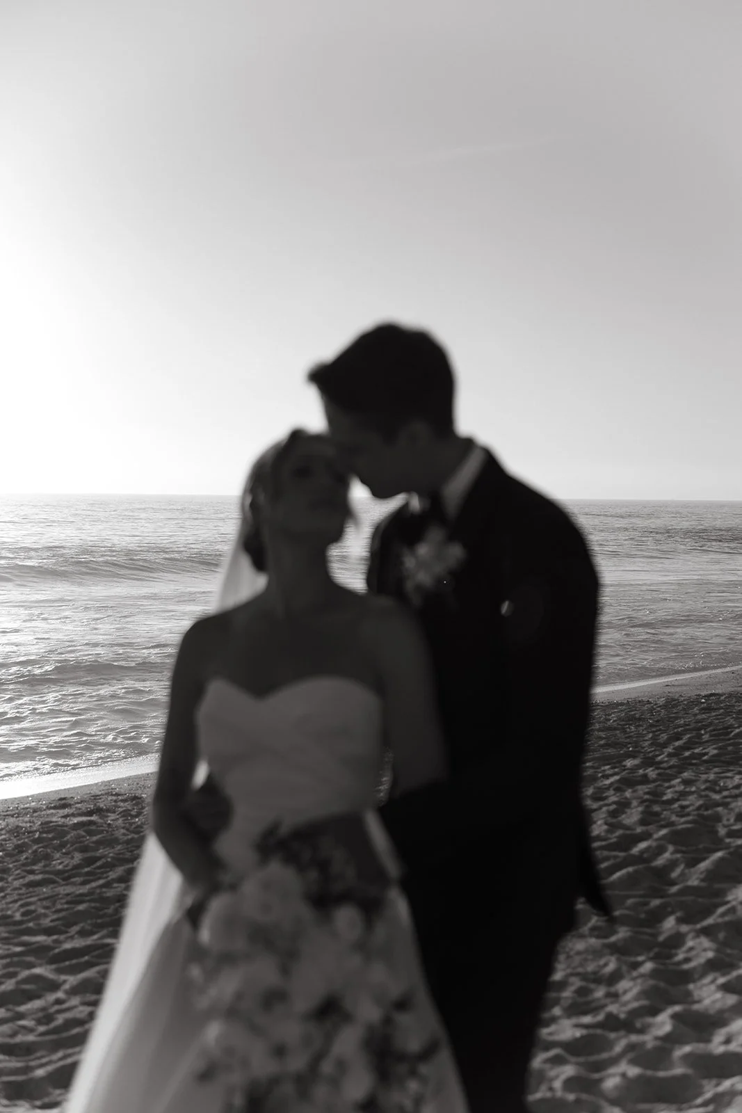 Soft-focus black and white portrait of the bride and groom embracing on the beach with waves and horizon in the background.