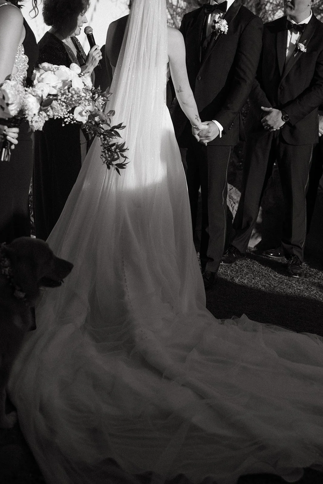 Black and white image of the couple holding hands during their vows at a timeless San Clemente Wedding Venue, highlighting the length of the bride’s veil and emotional moment.