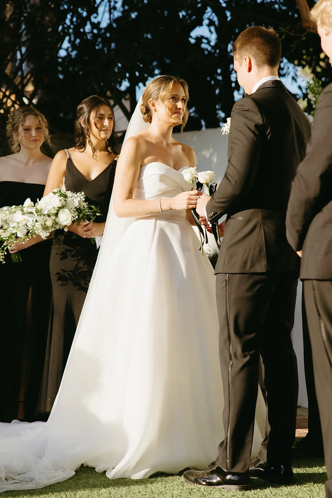 Close-up of the bride holding flowers as she faces the groom during the ceremony, with bridesmaids standing behind her in black dresses.