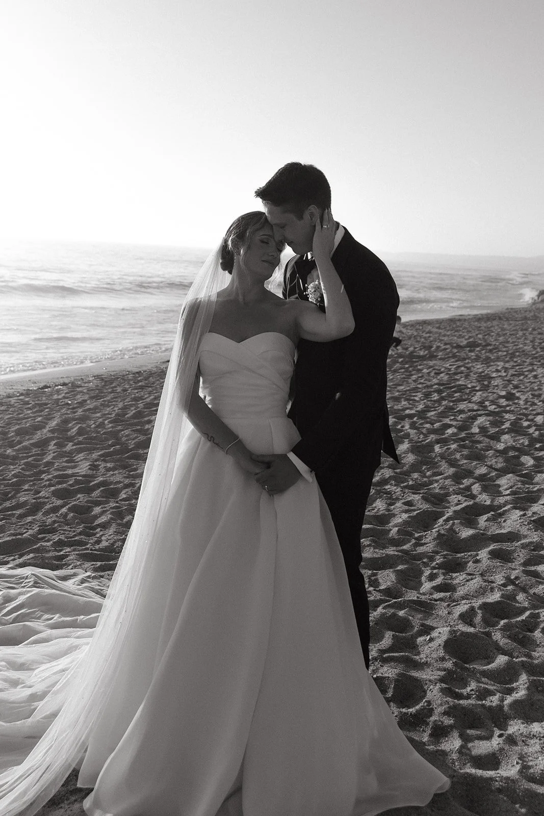 A black and white romantic sunset portrait of the bride and groom embracing on the beach near iconic San Clemente Wedding Venues, framed by soft waves and pastel sky tones.