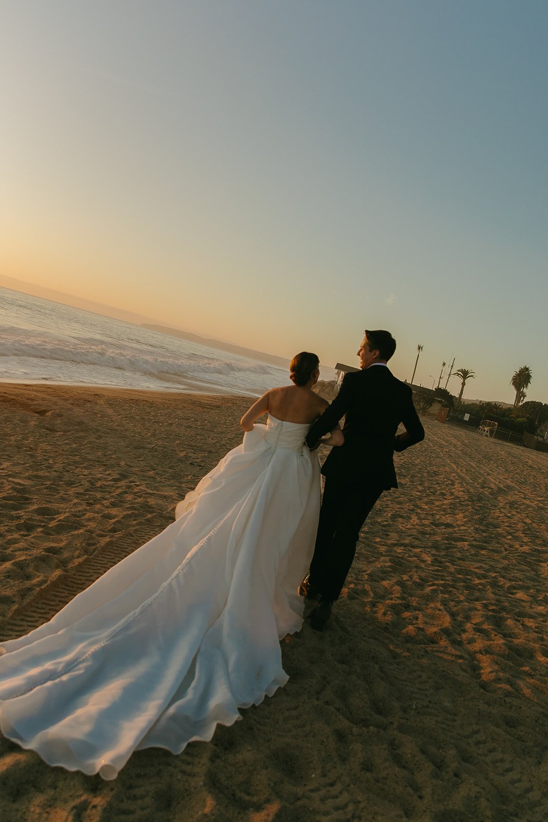 Bride and groom walking hand in hand along the shoreline after their ceremony, with palm trees and coastal homes in the distance.