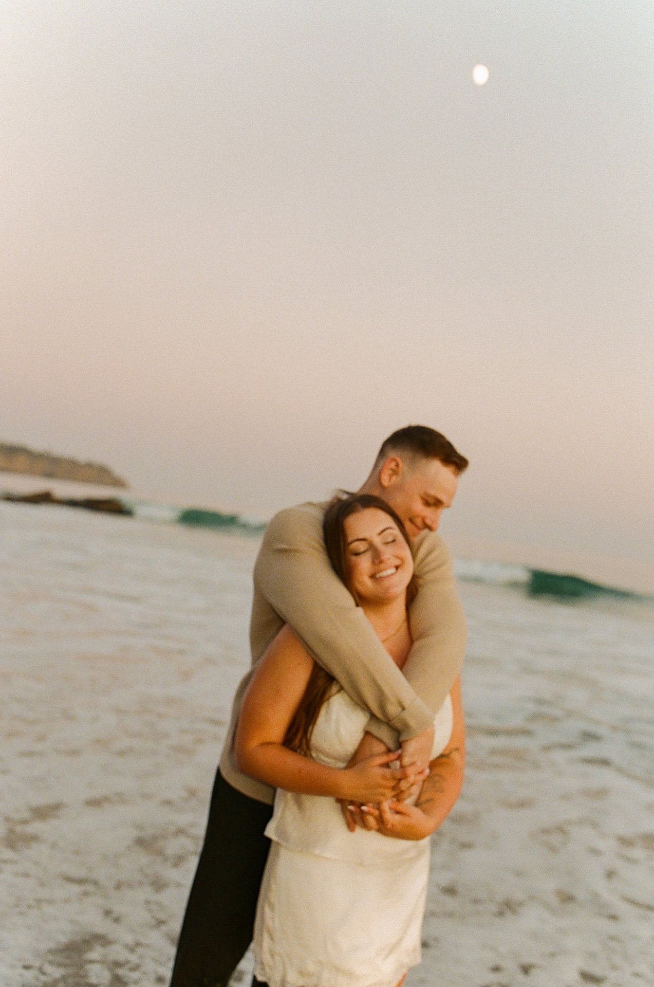 A couple embracing on the shoreline during sunset, with waves rolling in and the moon visible in the pastel sky.
