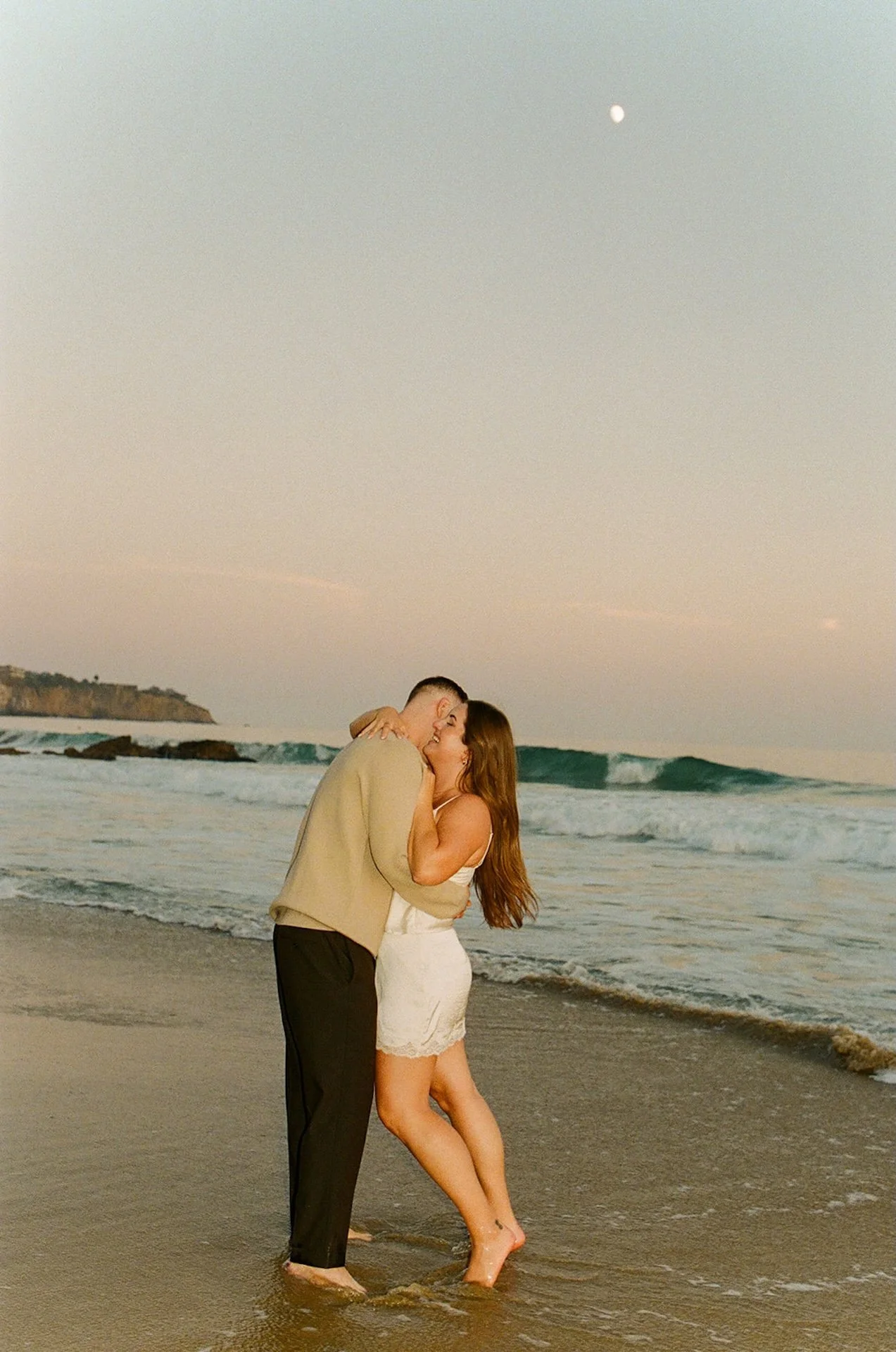 Beach Engagement Photo Ideas of a couple kissing in the shallow surf with coastal cliffs in the background and soft golden-hour lighting.