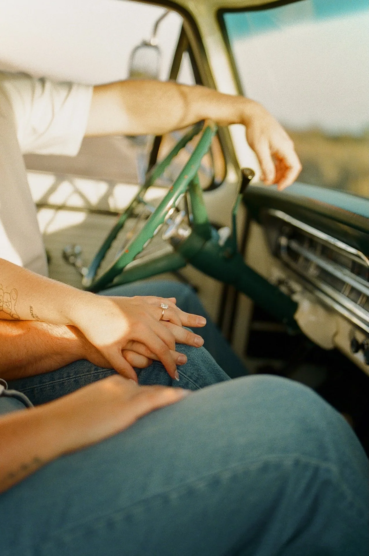 Beach Engagement Photo Ideas showing intertwined hands on denim-clad legs inside a classic truck, with sunlight casting soft shadows across the interior.