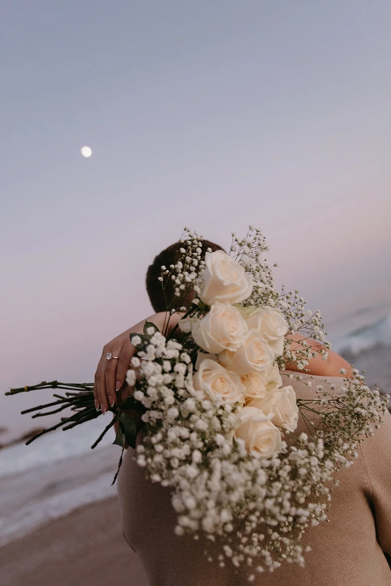 A romantic beach portrait of a bouquet of white roses and baby’s breath held over a couple’s embrace, with the moon and ocean in the background at dusk.