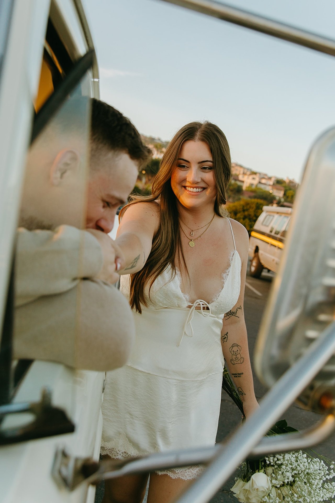 A candid moment of a woman smiling as her fiance kisses her hand through the truck window, with a bouquet of white flowers resting at her side.