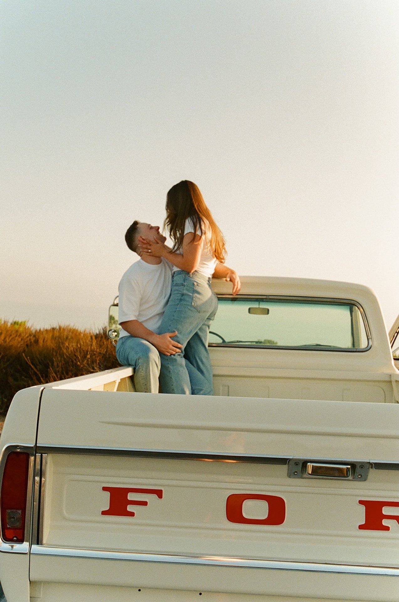 A wide shot of a couple standing in the bed of a classic truck, embracing and kissing with soft golden-hour light and an open sky backdrop.