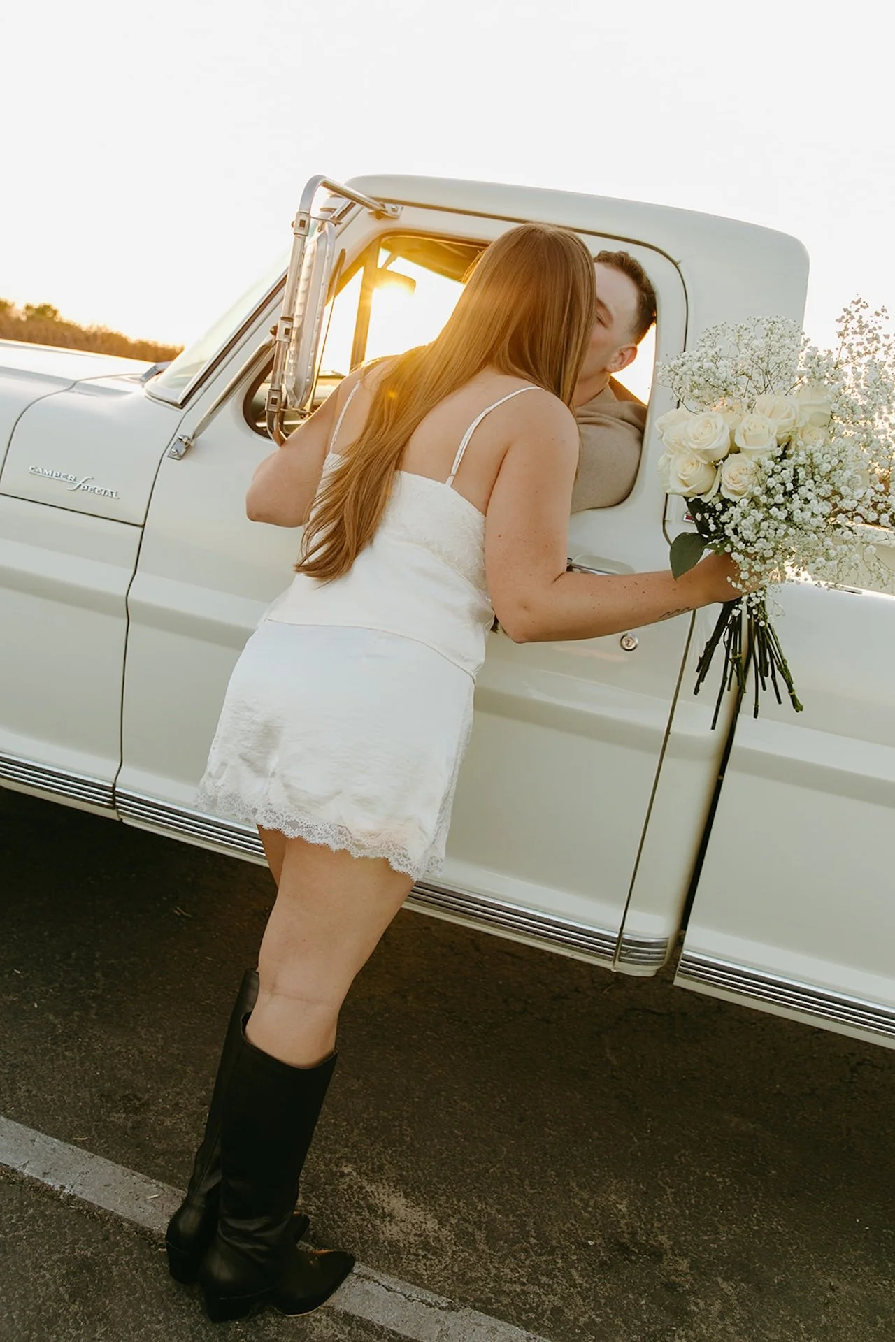 Beach Engagement Photo Ideas showing a woman in a white dress leaning into a truck window to kiss her partner, holding a bouquet of white roses and baby’s breath in warm sunset light.