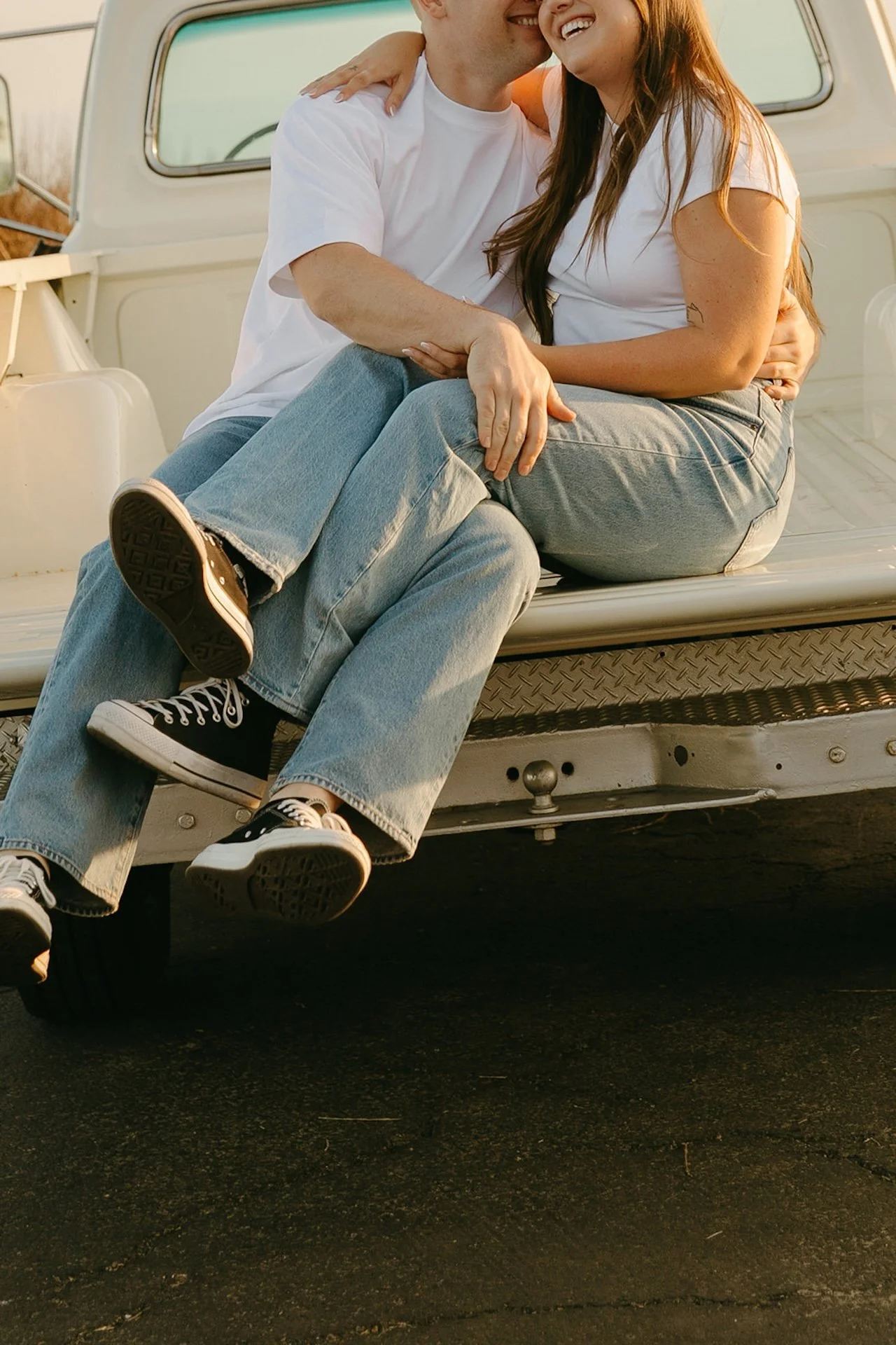 A close-up of a couple sitting on the back of a vintage pickup truck, laughing together with their legs crossed and soft sunset light hitting their faces.