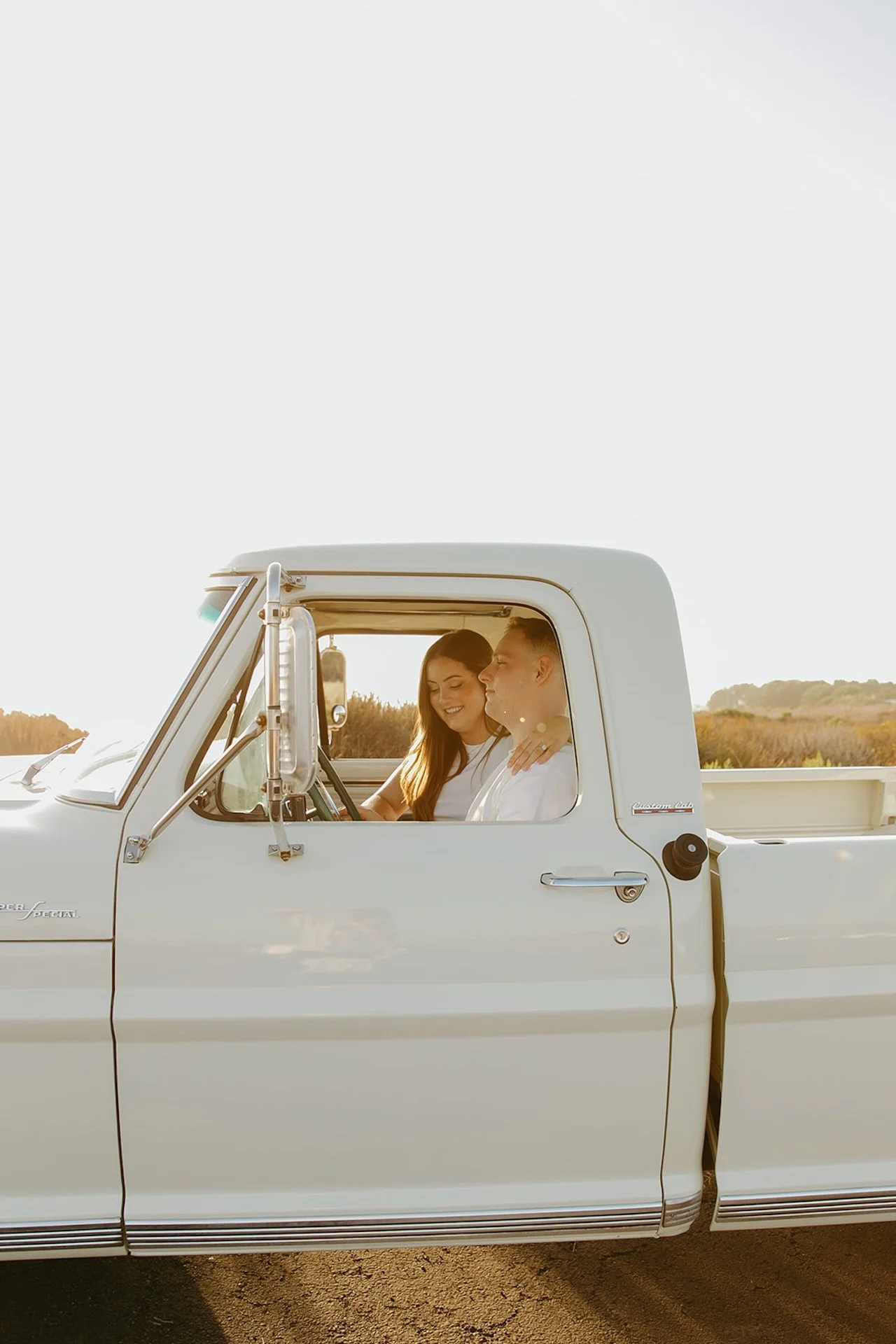 Beach Engagement Photo Ideas of a couple sitting inside a vintage white truck during golden hour, smiling and leaning into each other in relaxed white tops and denim.