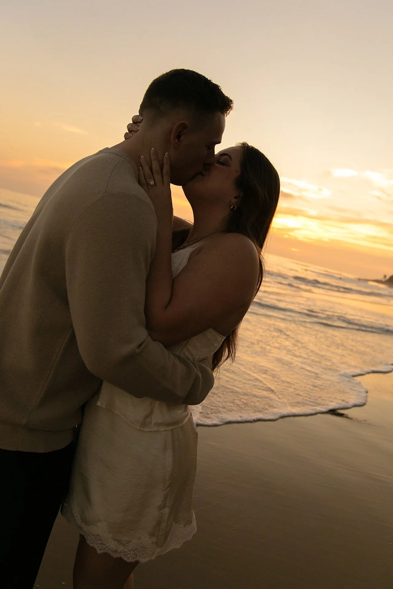 A silhouette-style beach portrait of a couple kissing at sunset with waves and warm golden sky creating a romantic backdrop.