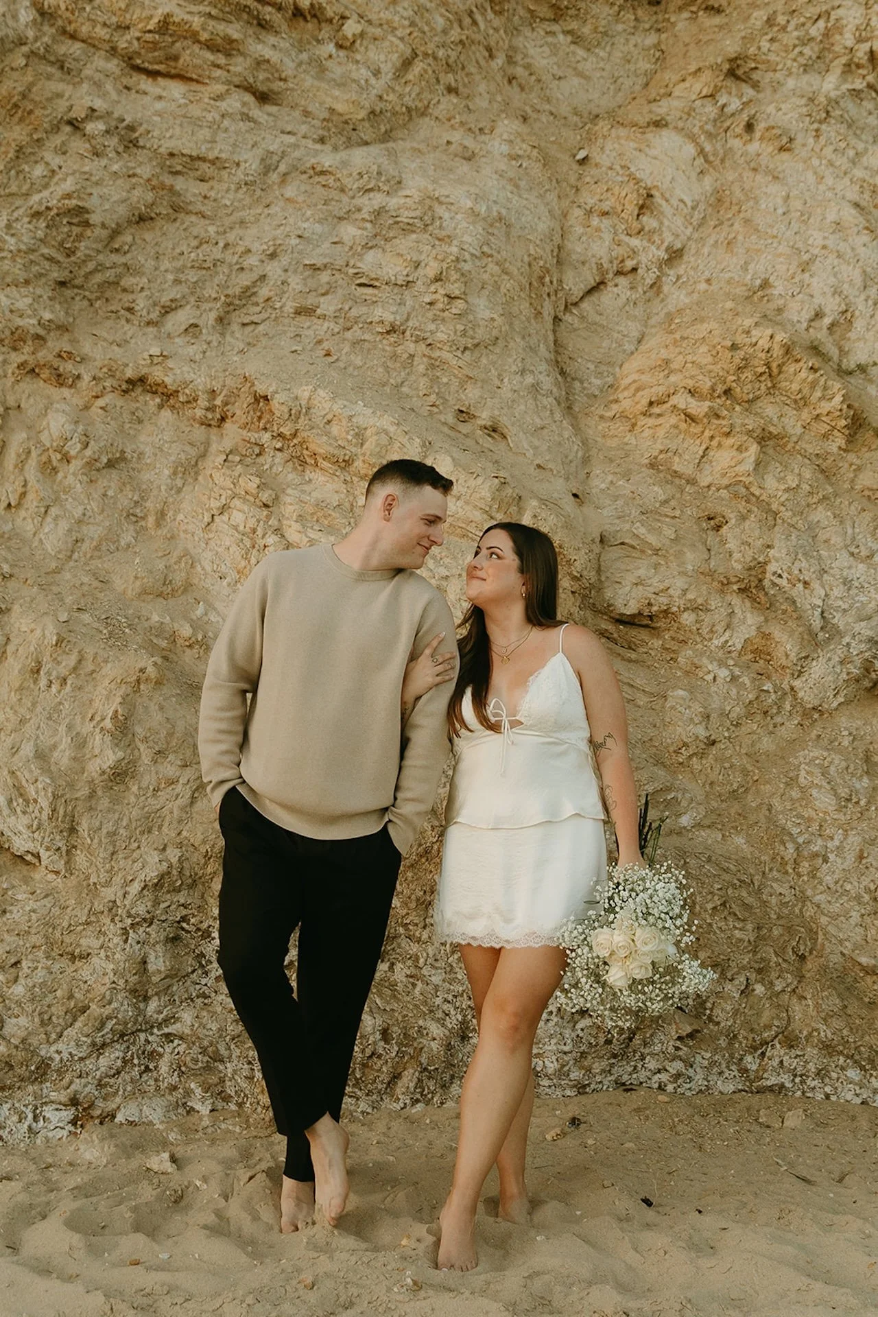 A couple standing against textured coastal cliffs, holding a bouquet of white roses and baby’s breath while looking into each other’s eyes.