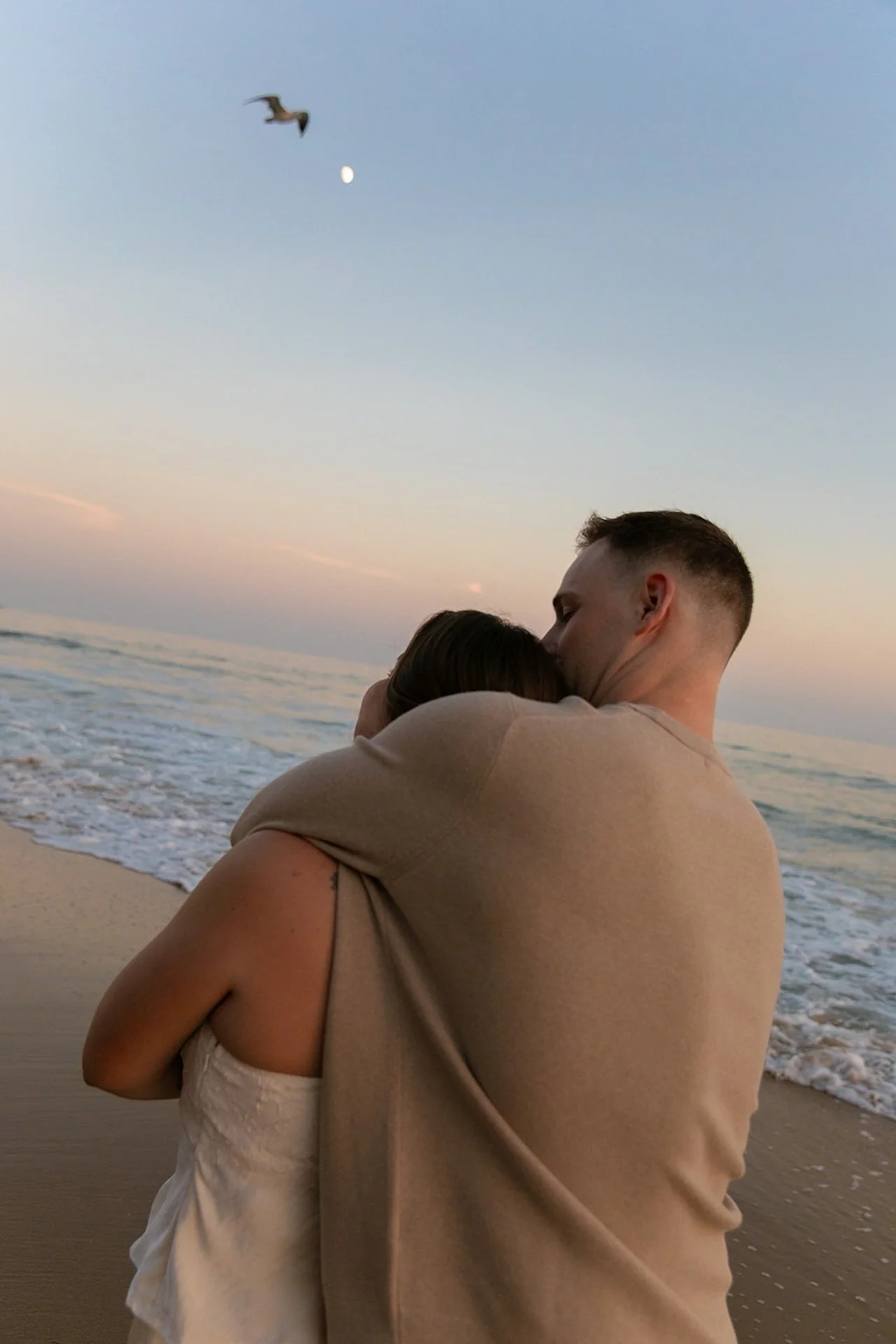 Beach Engagement Photo Ideas of a couple embracing while standing in the shallow surf, with gentle waves and a soft sunset horizon behind them.
