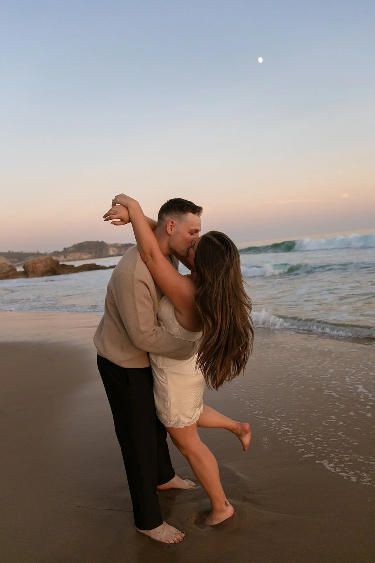 A couple kissing on the beach with the woman lifting one leg and wrapping her arms around her partner during golden hour.