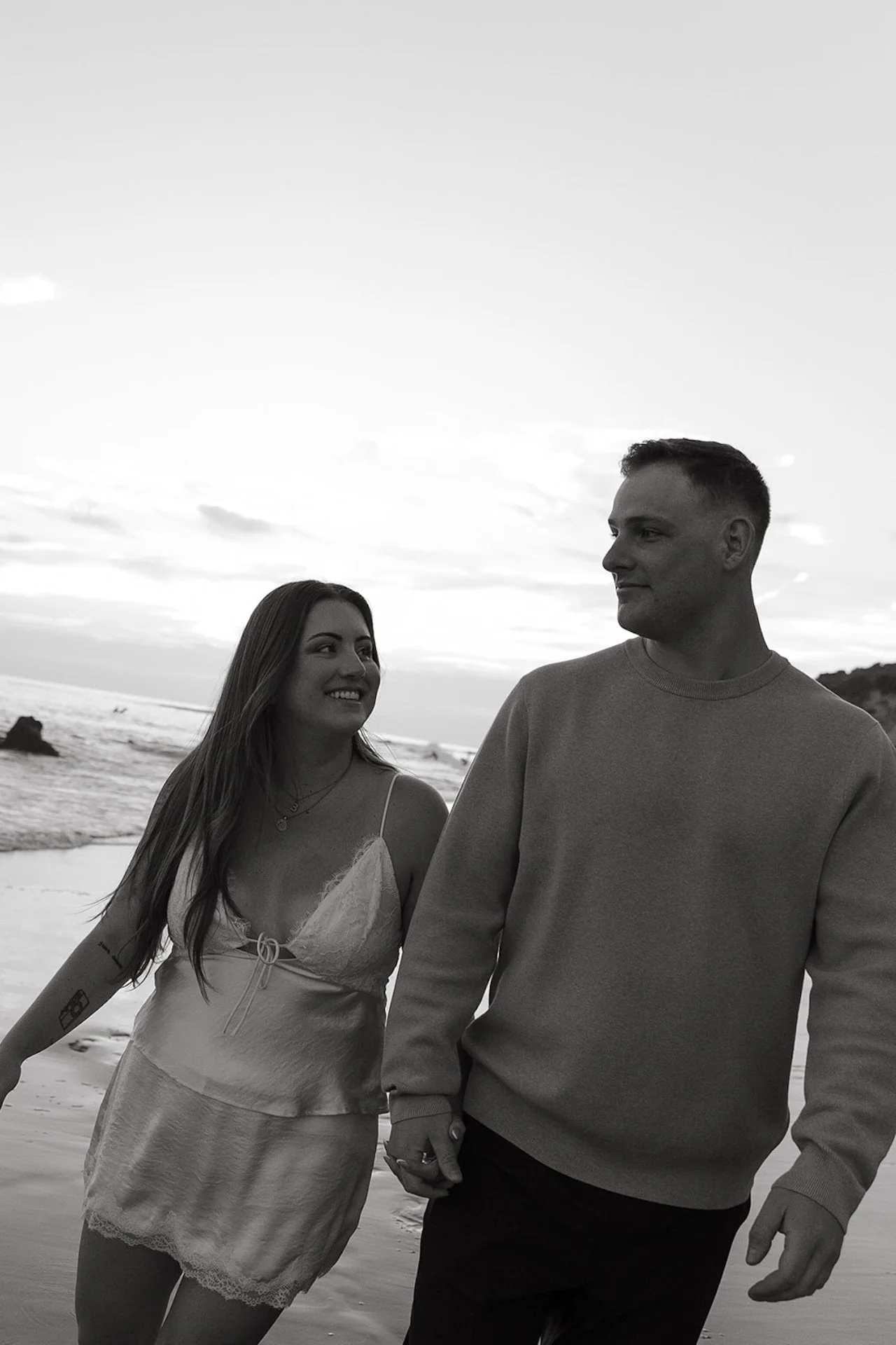 Beach Engagement Photo Ideas showing a couple walking barefoot on the beach, holding hands and smiling with coastal cliffs and waves in the background.